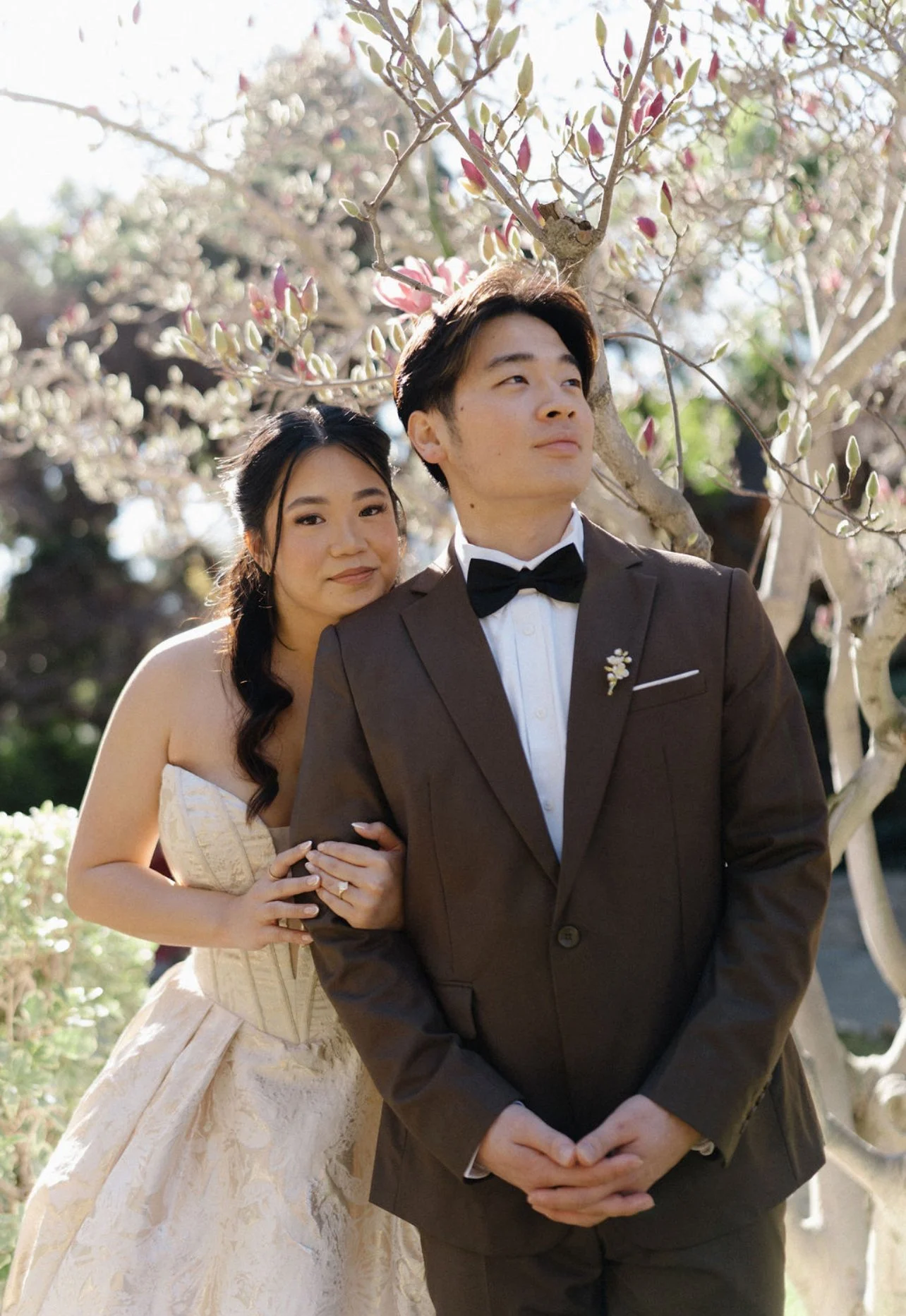 A couple dressed in wedding attire standing outdoors under a flowering tree, with the woman resting her head on the man's shoulder.