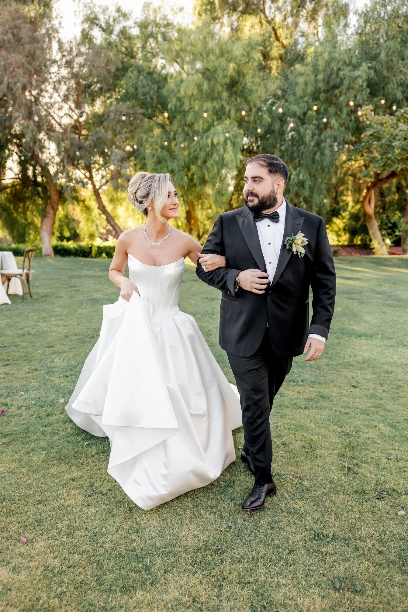 A bride and groom walking arm-in-arm outdoors on a sunny day, with trees and string lights in the background. The bride is wearing a strapless white wedding gown and a pearl necklace. The groom is dressed in a black tuxedo with a bow tie and a bouton