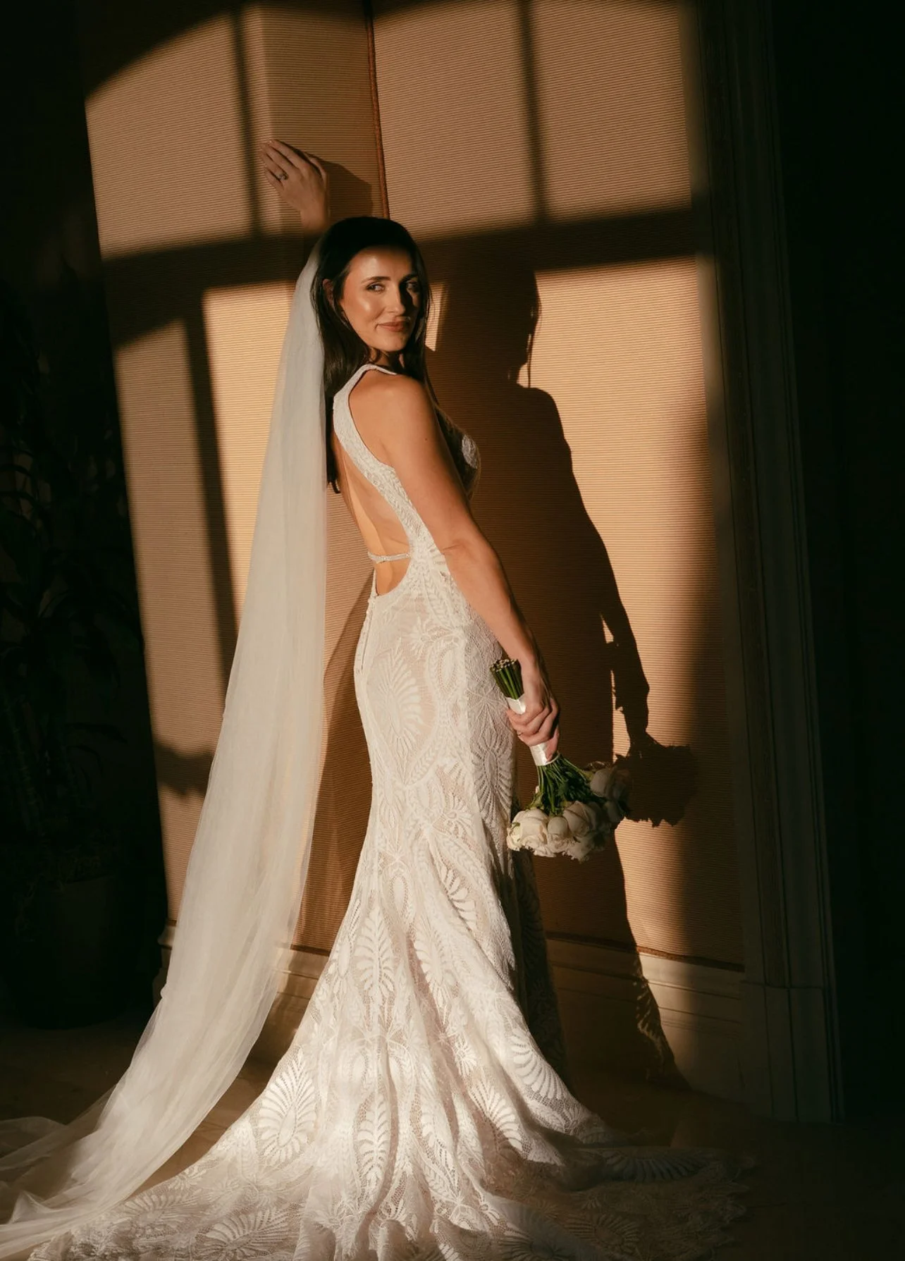 A bride in a detailed lace wedding gown holding a bouquet of white roses, standing by a window with her shadow cast on the wall behind her.