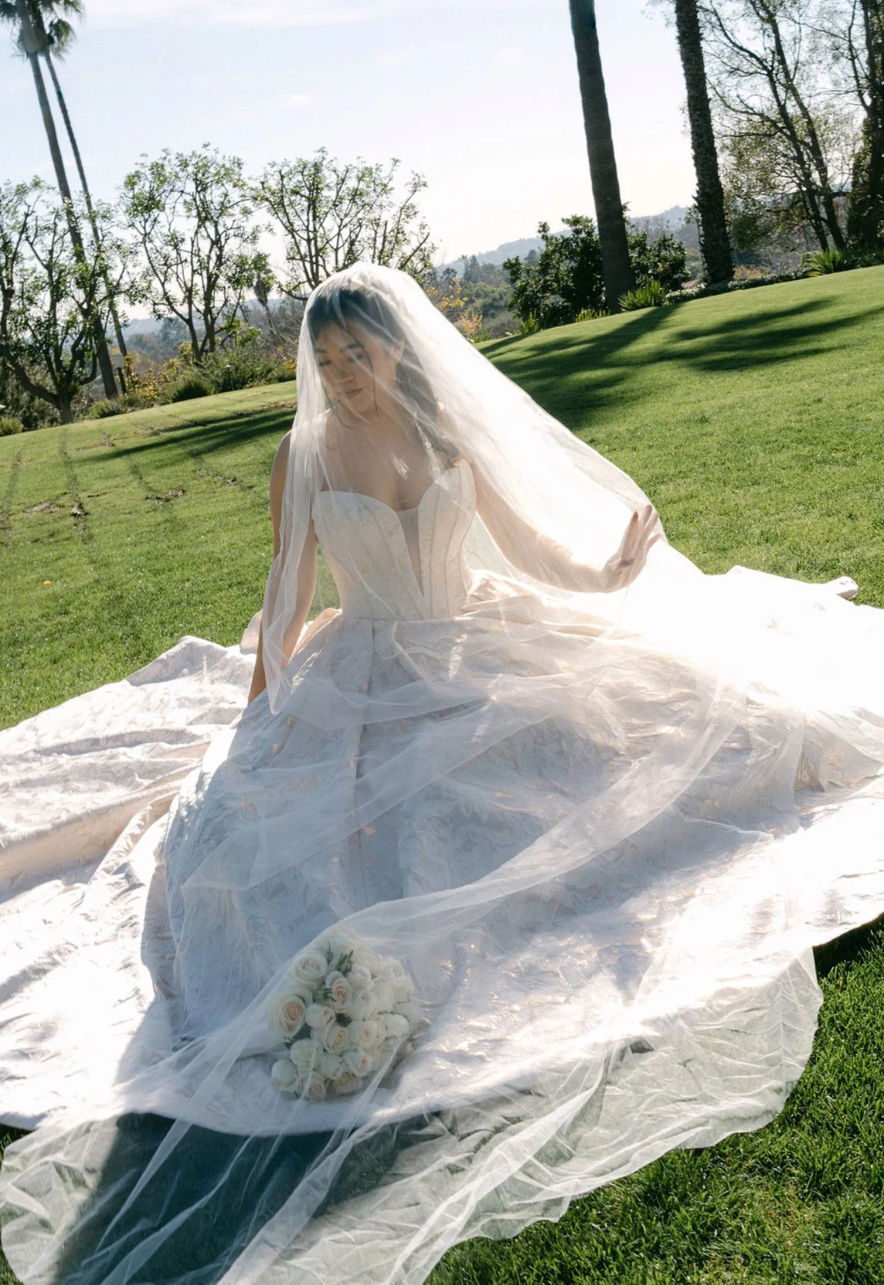 A woman in a wedding dress sitting on grass with a veil covering her face, outdoors with trees and blue sky in the background. A bouquet of white roses is placed in front of her.