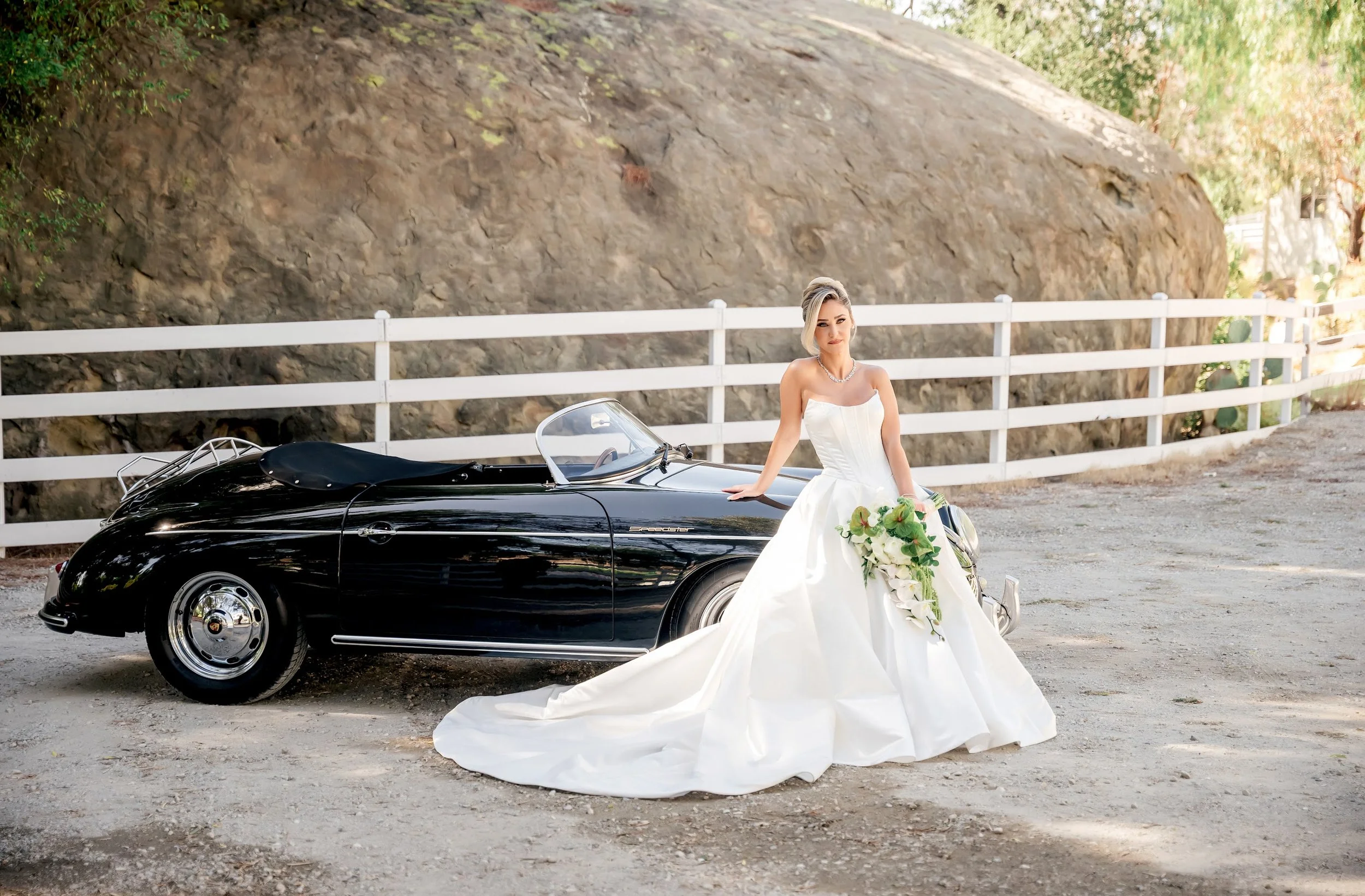 A woman in a white wedding dress standing next to a black vintage sports car, holding a bouquet of white flowers, outdoors with a large rocky hill and wooden fence in the background.