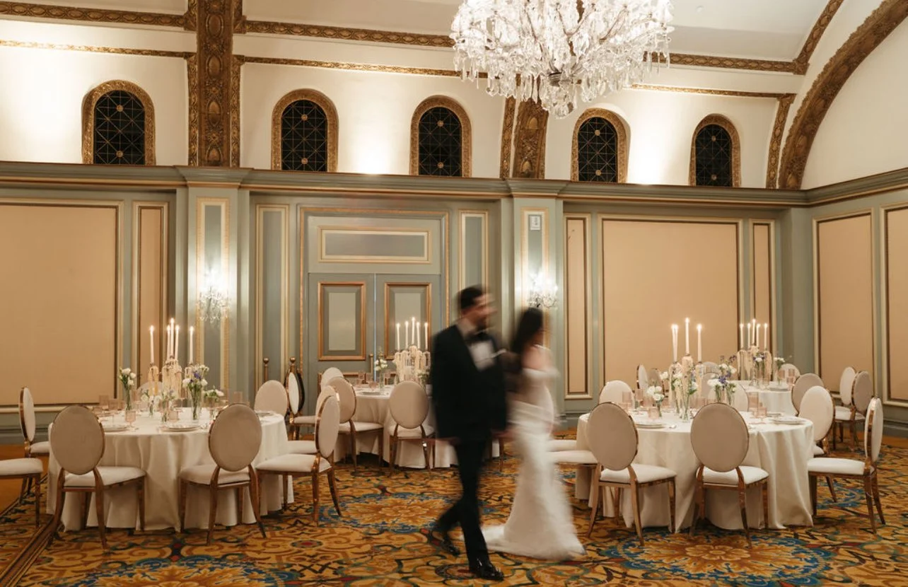 Elegant banquet hall decorated with round tables with white tablecloths, floral centerpieces, and candelabras, with a chandeliers hanging overhead. A couple is walking in the background, possibly at a wedding reception.