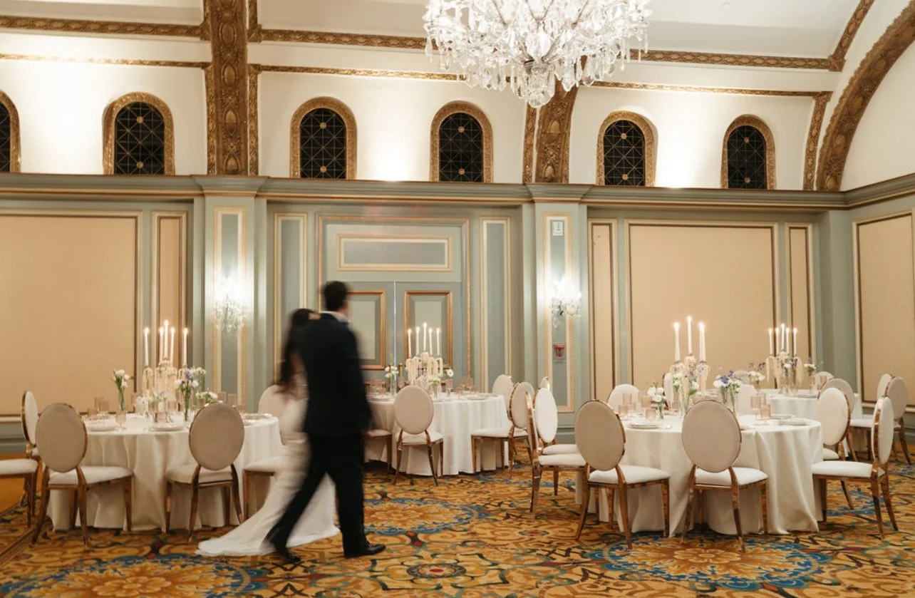 Elegant banquet hall with round tables, white tablecloths, floral centerpieces, candles, ornate chairs, and a large crystal chandelier, with a couple in wedding attire walking in.