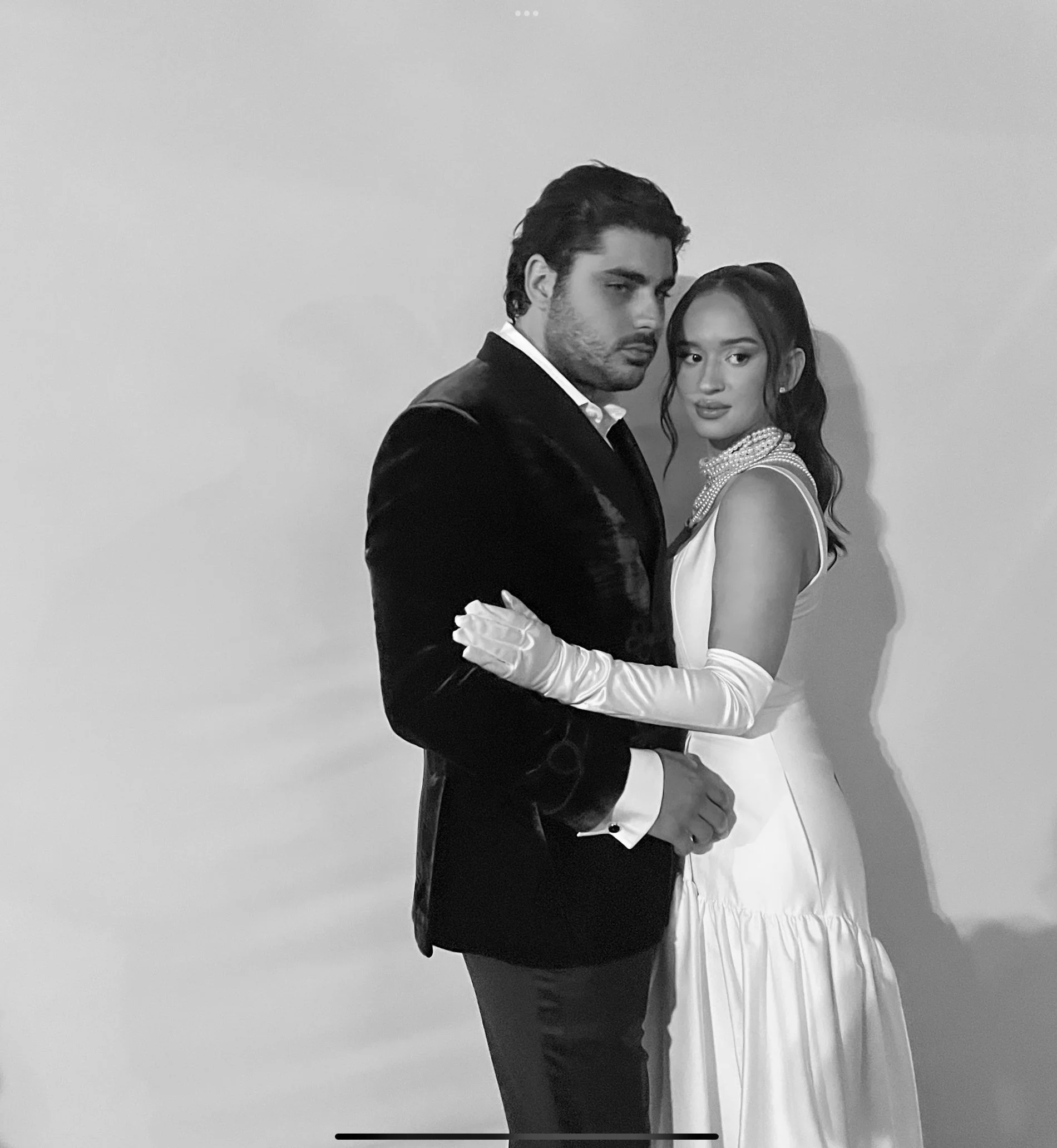 A black and white portrait of a man and woman dressed in formal attire, standing closely together against a plain background.