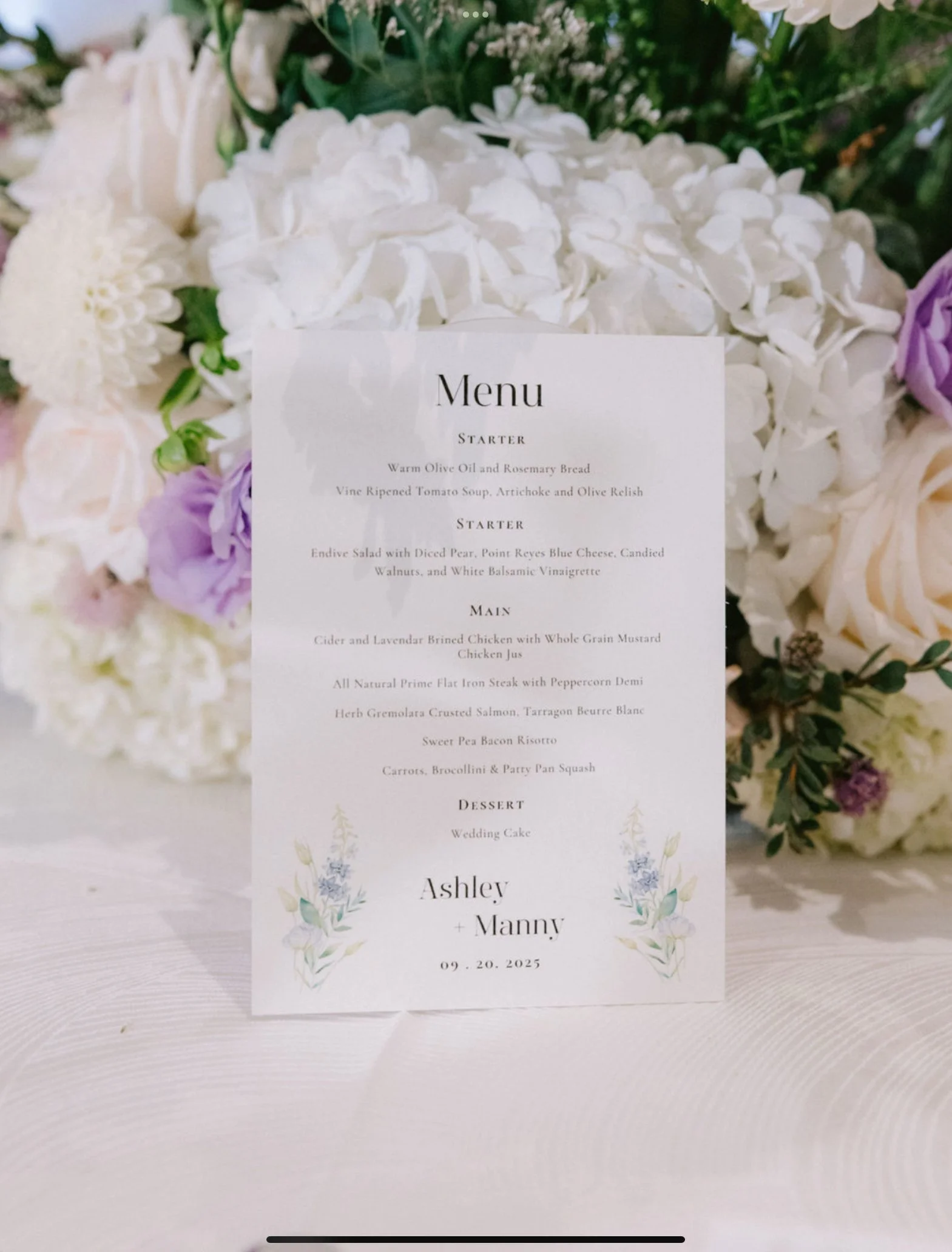 Wedding menu card placed on a white floral backdrop with white and purple flowers surrounding it. The menu includes starters, main courses, dessert, and the wedding date.