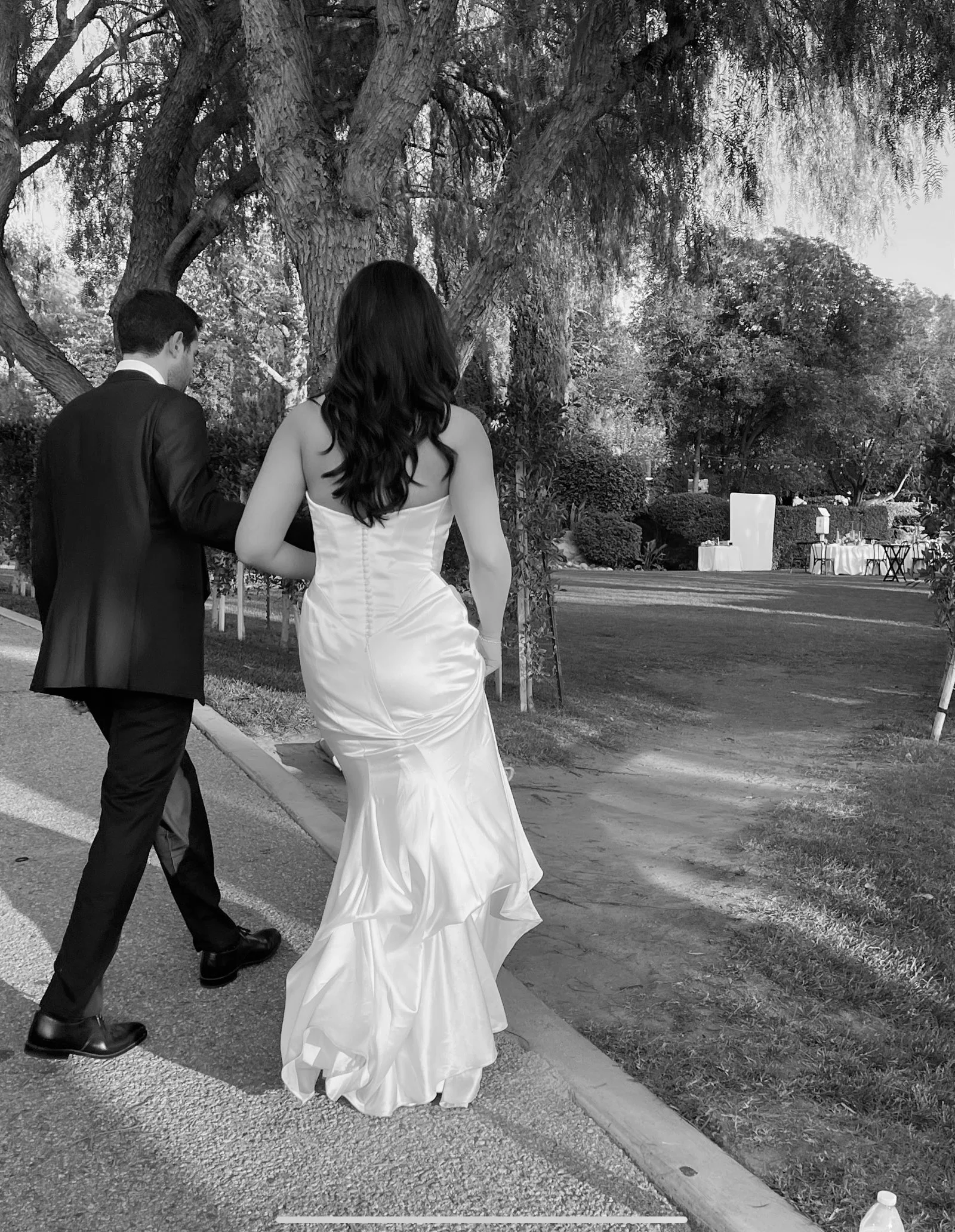 A black and white photo of a bride and groom walking outdoors near trees and bushes. The bride is in a strapless wedding dress with long, dark hair, and the groom is in a dark suit with short dark hair.