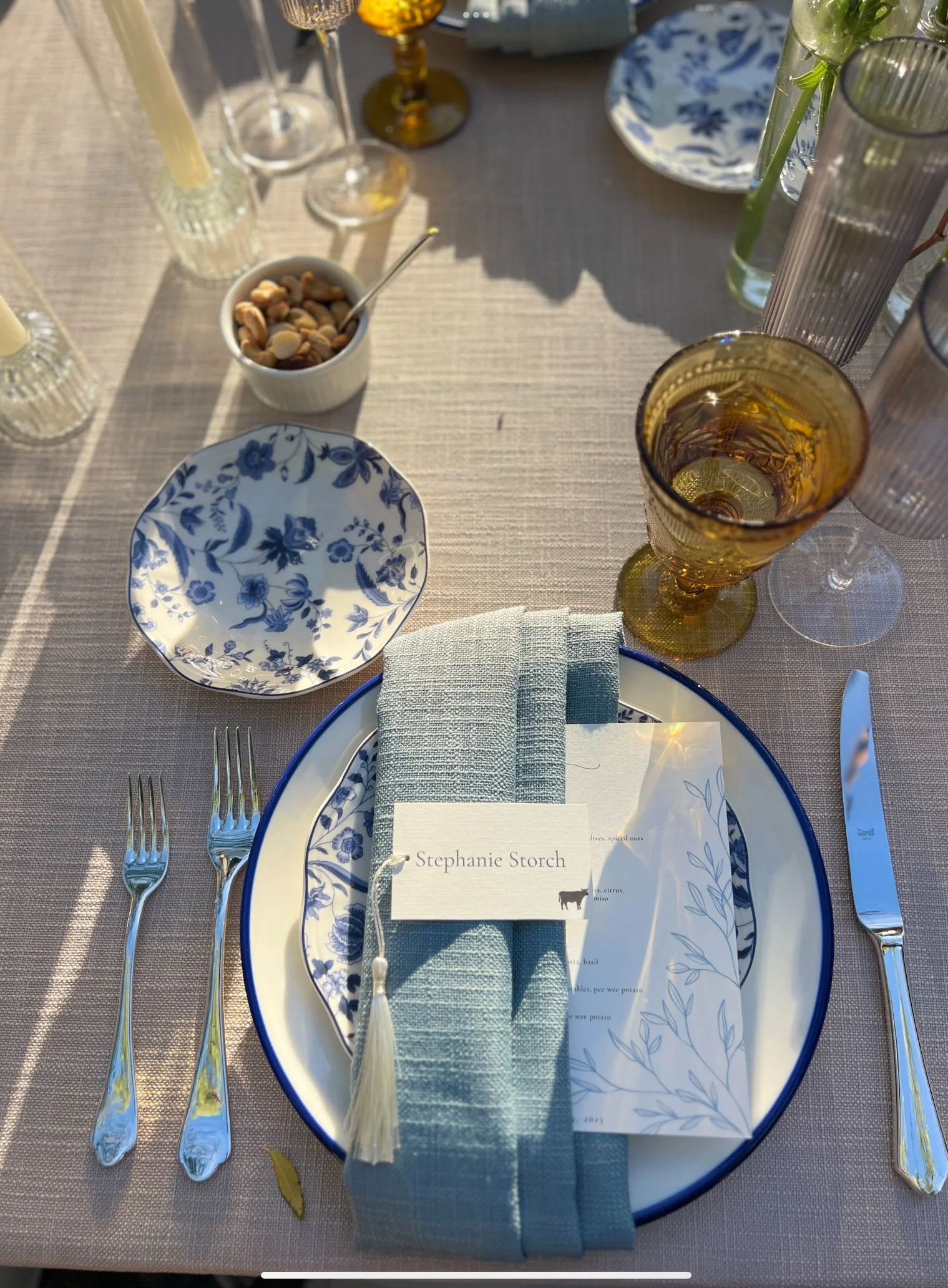 Table setting with a blue-rimmed white plate, three forks, a knife, a blue napkin, and a menu card with the name 'Stephanie Storch'. There are several glasses, a small bowl of mixed nuts, a menu featuring various dishes, and some vases with tall gree