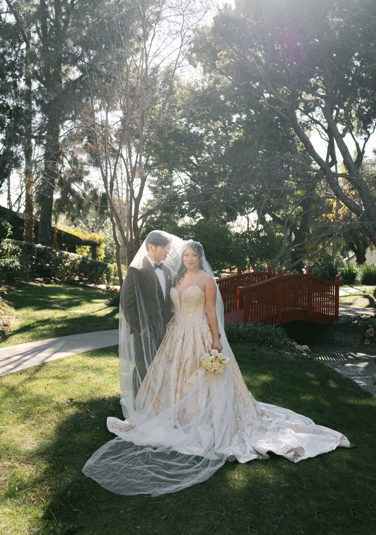 A bride and groom standing together outdoors on a sunny day, with trees and a small red bridge in the background. The bride is holding a bouquet and wearing a wedding gown with a long train and veil. The groom is dressed in a tuxedo.