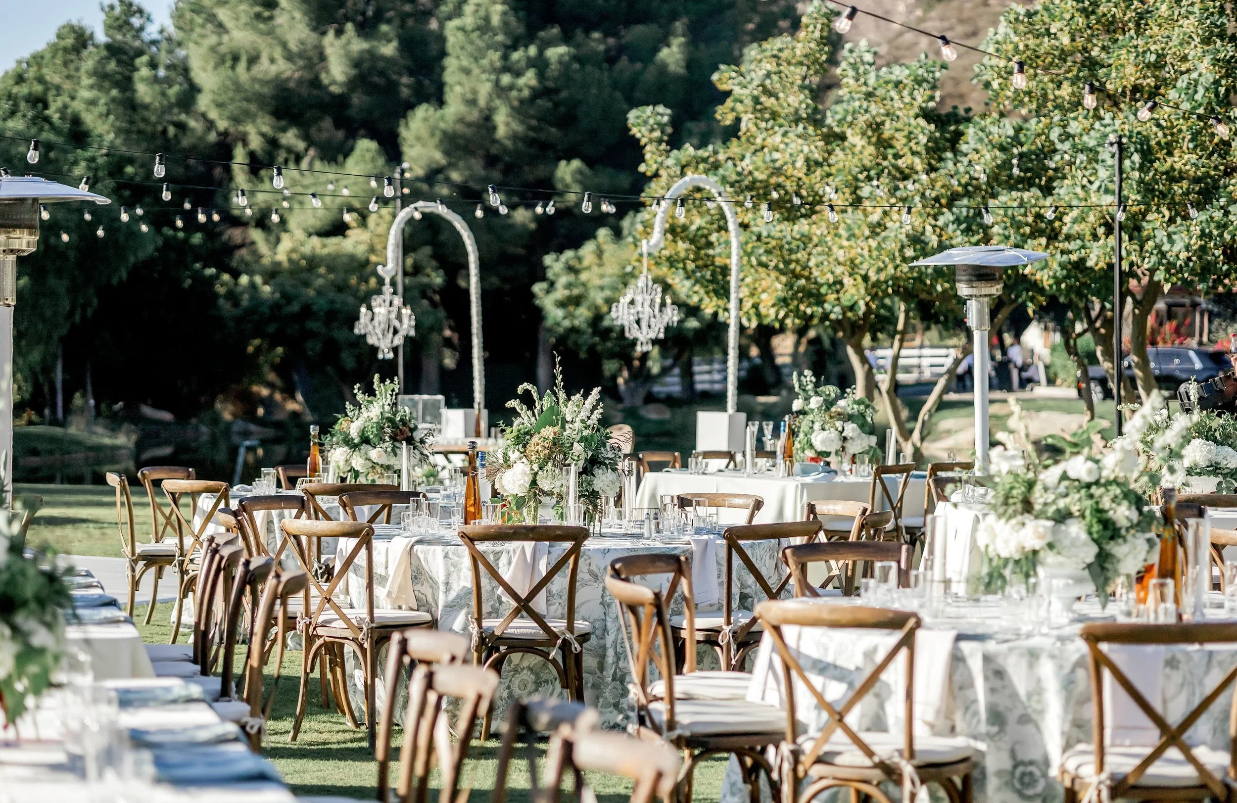 Outdoor wedding reception setup with tables decorated with flowers and surrounded by chairs, string lights hanging above, and trees in the background.