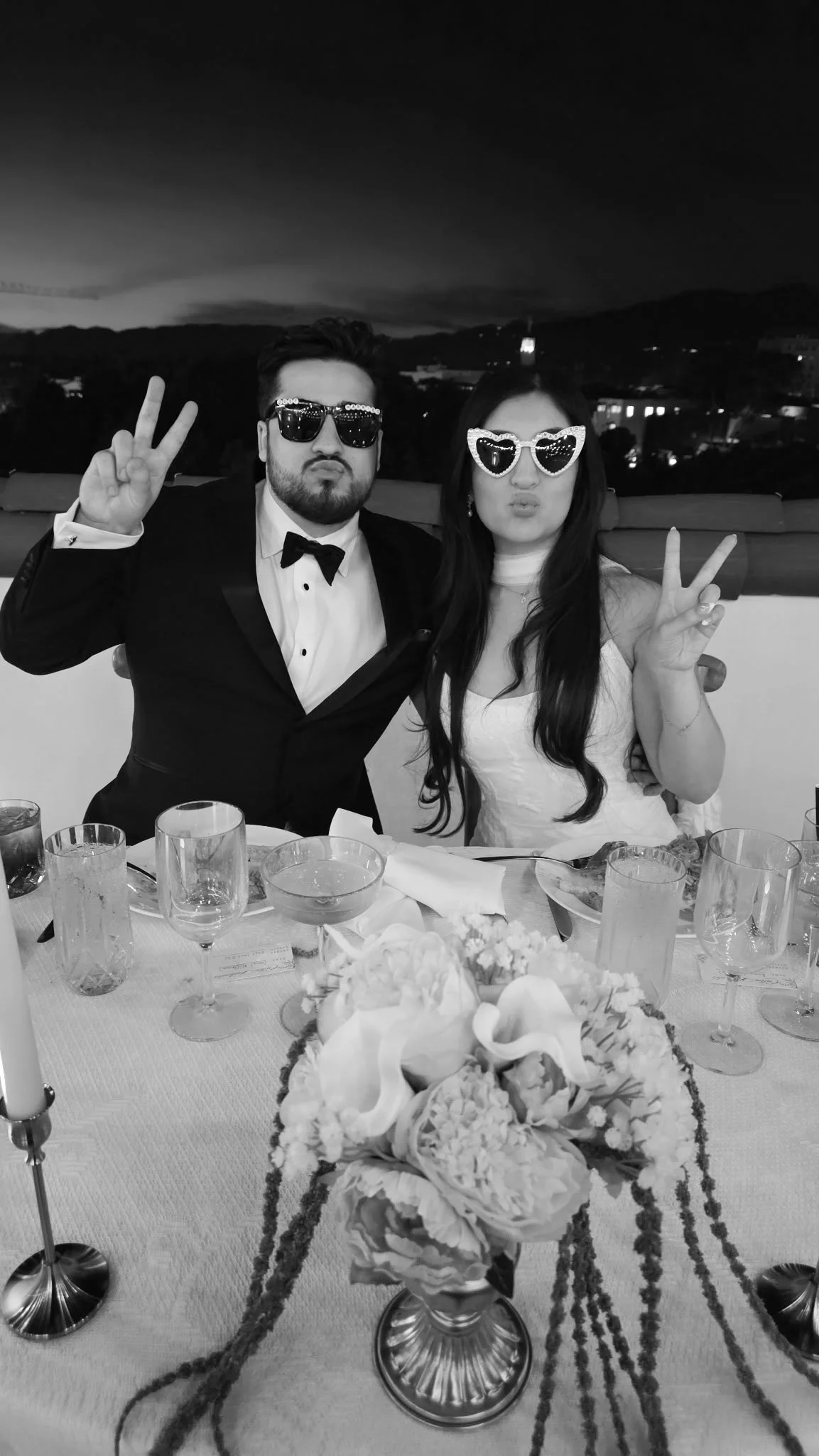 A black-and-white photo of a man and woman at a formal dining event. The man is wearing a tuxedo with a bow tie, sunglasses, and making a peace sign. The woman is wearing a white dress, heart-shaped sunglasses, and also making a peace sign. They are 