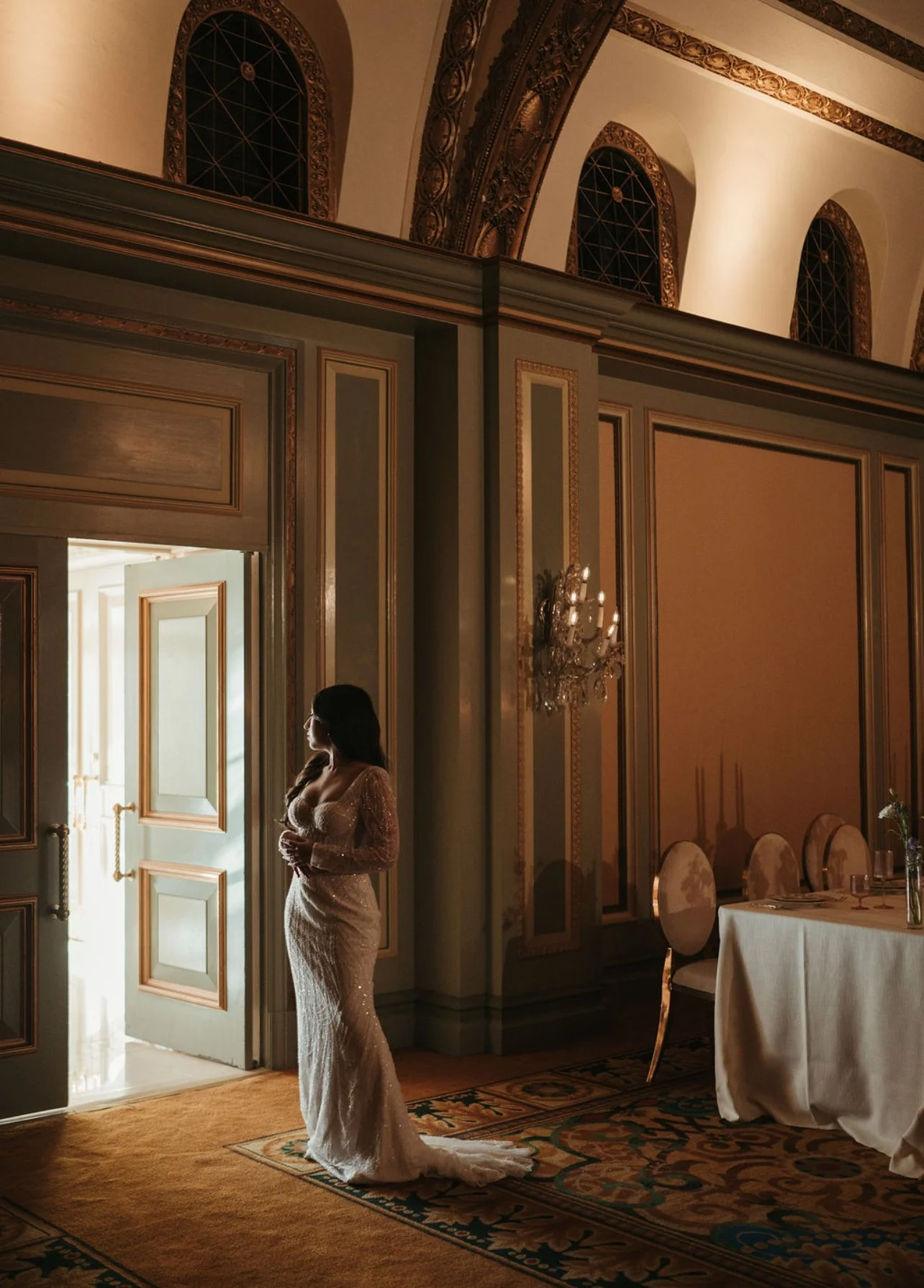 A woman in a white gown standing in a luxurious, ornate room with gold accents, looking out of an open door.