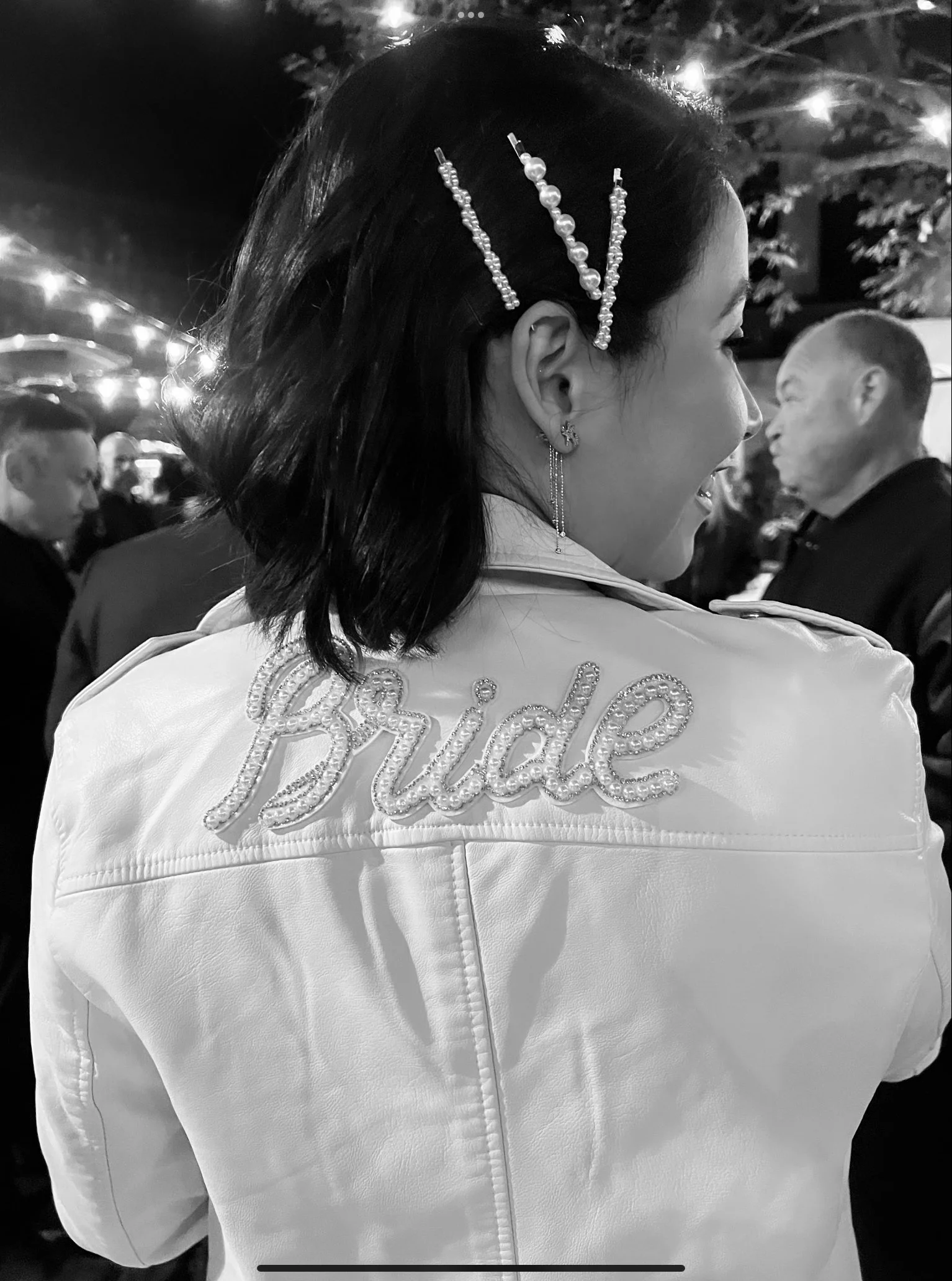 A woman with dark hair styled with pearl and beaded hairpins, wearing earrings and a white jacket with shiny embroidery, standing in a dimly lit outdoor setting with string lights and trees, with other people in the background.