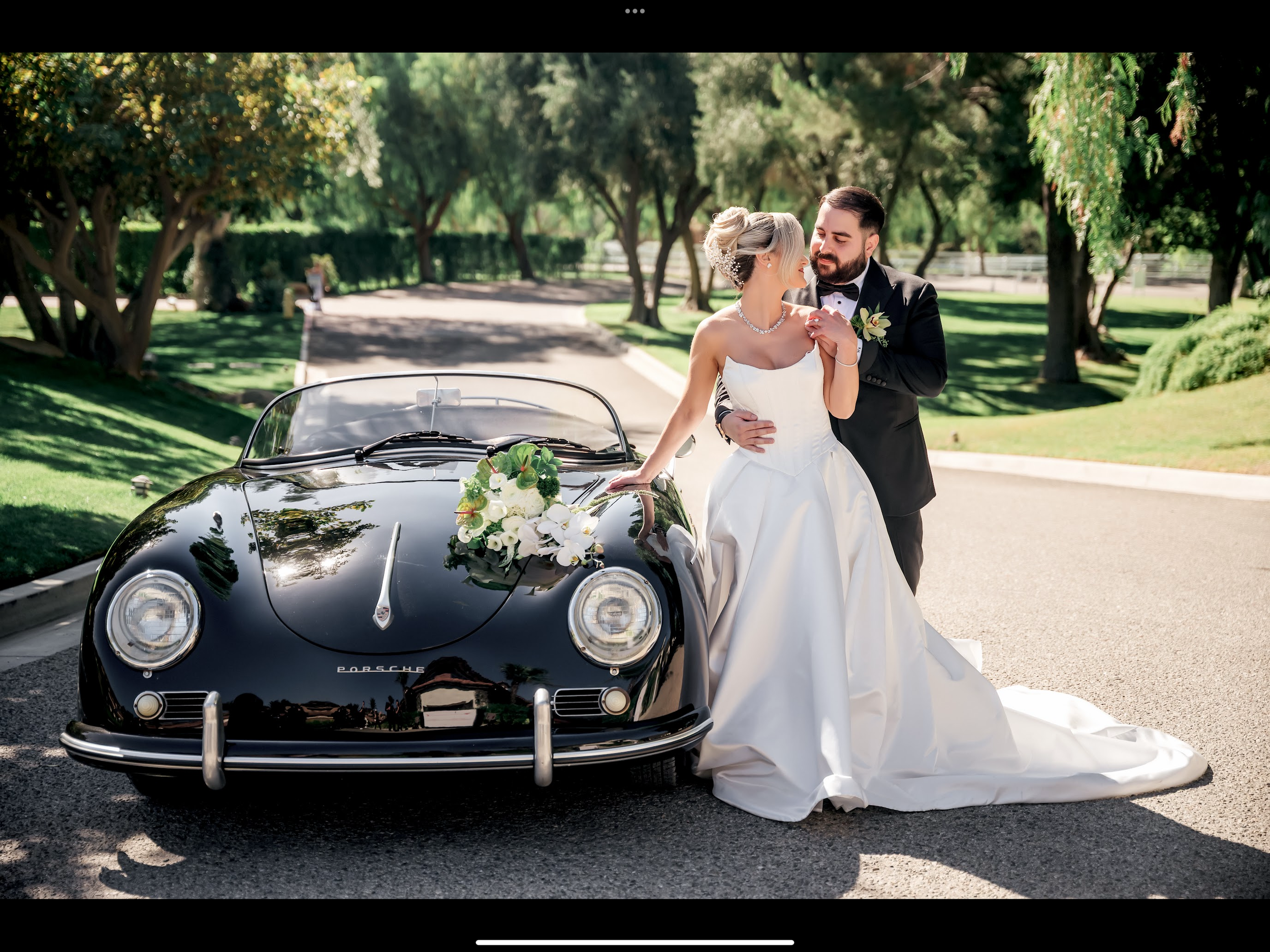 A bride and groom sharing an intimate moment next to a vintage black Porsche convertible decorated with wedding flowers in a park-like setting.