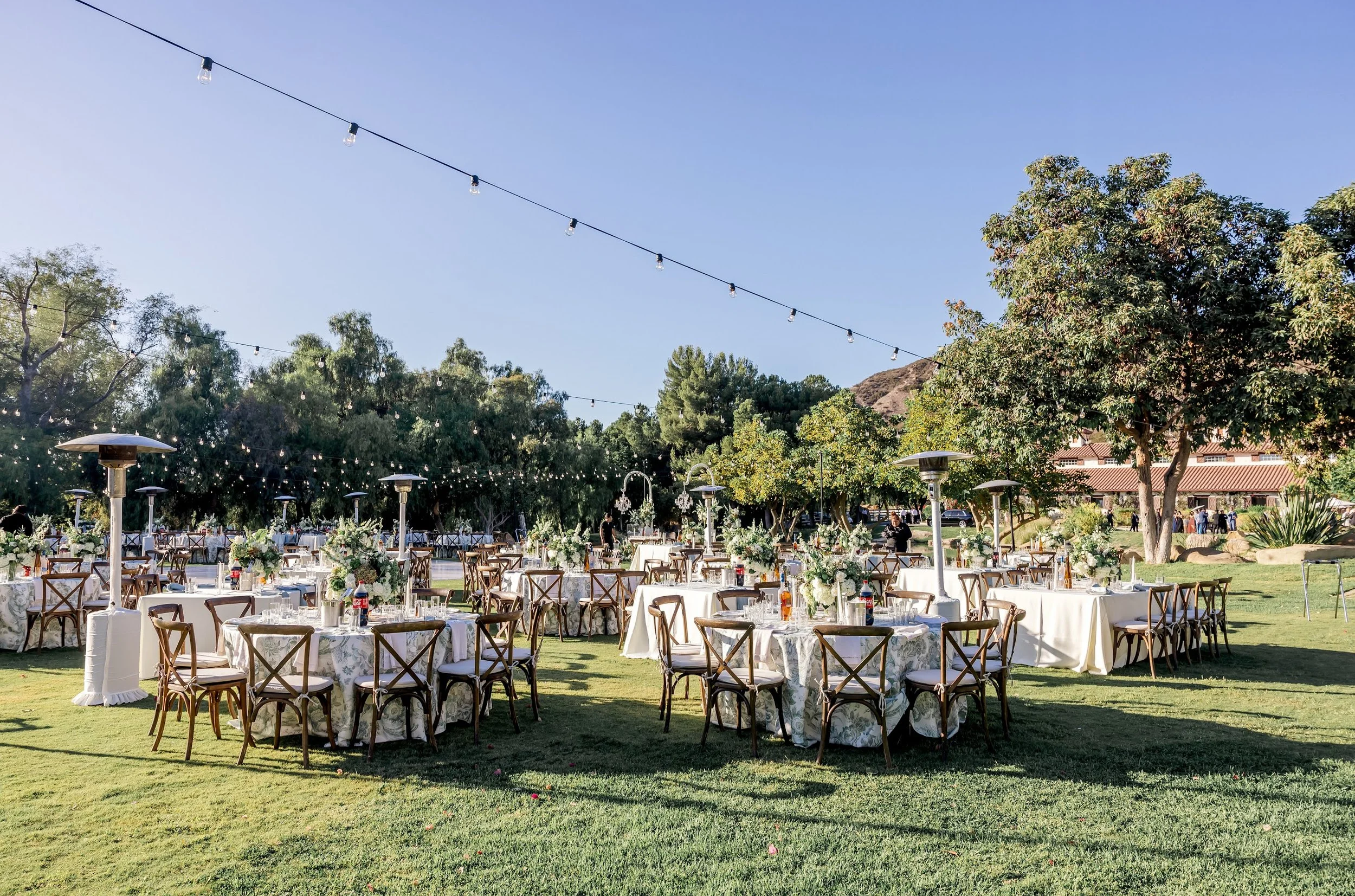 An outdoor wedding reception area with round tables draped in floral tablecloths, surrounded by wooden chairs, set up on grass with trees and string lights overhead against a clear blue sky.