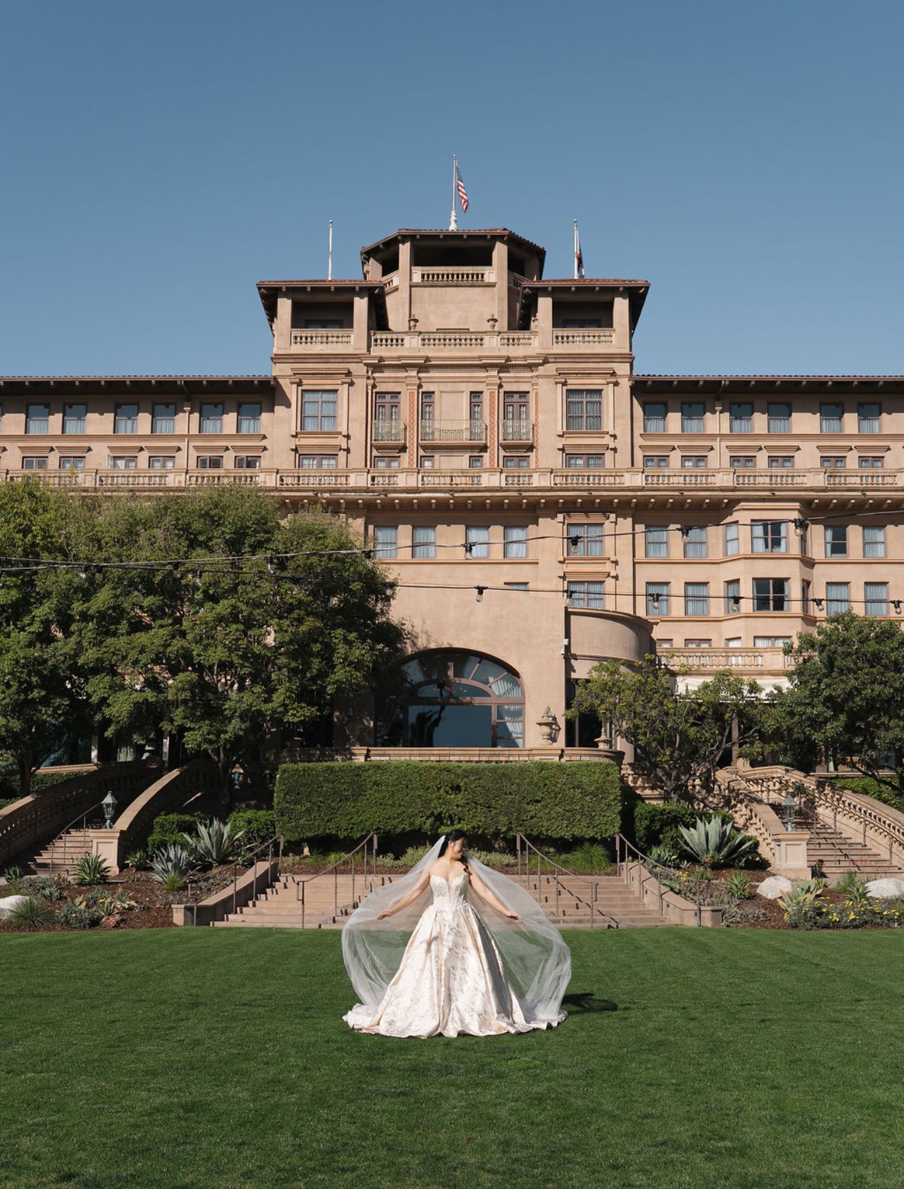 A bride in a wedding gown and veil stands on a well-manicured lawn, with a large historic building with multiple stories and flags in the background. The sky is clear and blue.