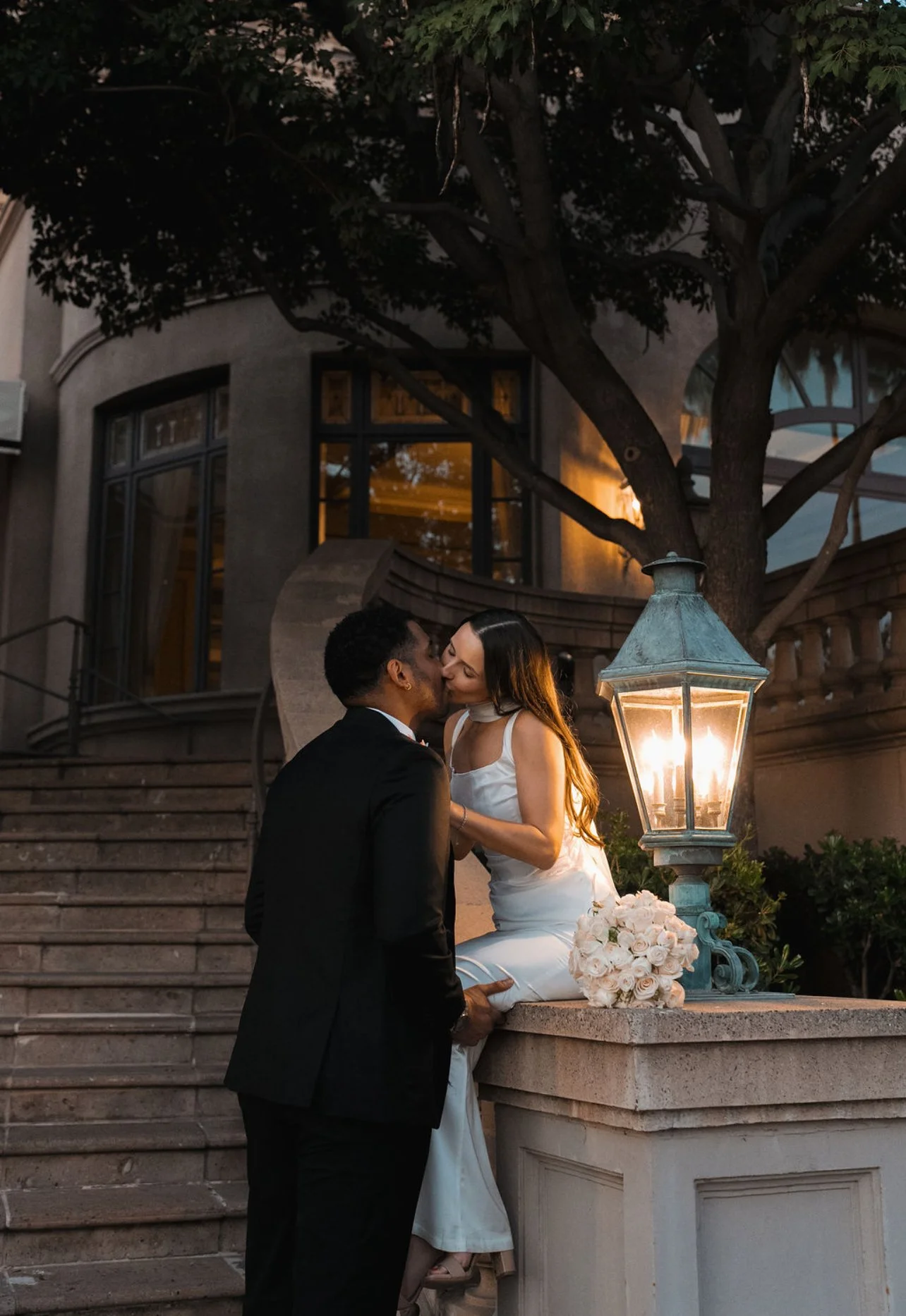 A bride and groom sharing a kiss outdoors near a streetlamp on the steps of a building at dusk. The bride is sitting on the ledge with a bouquet of white roses, and the groom is standing in front of her.