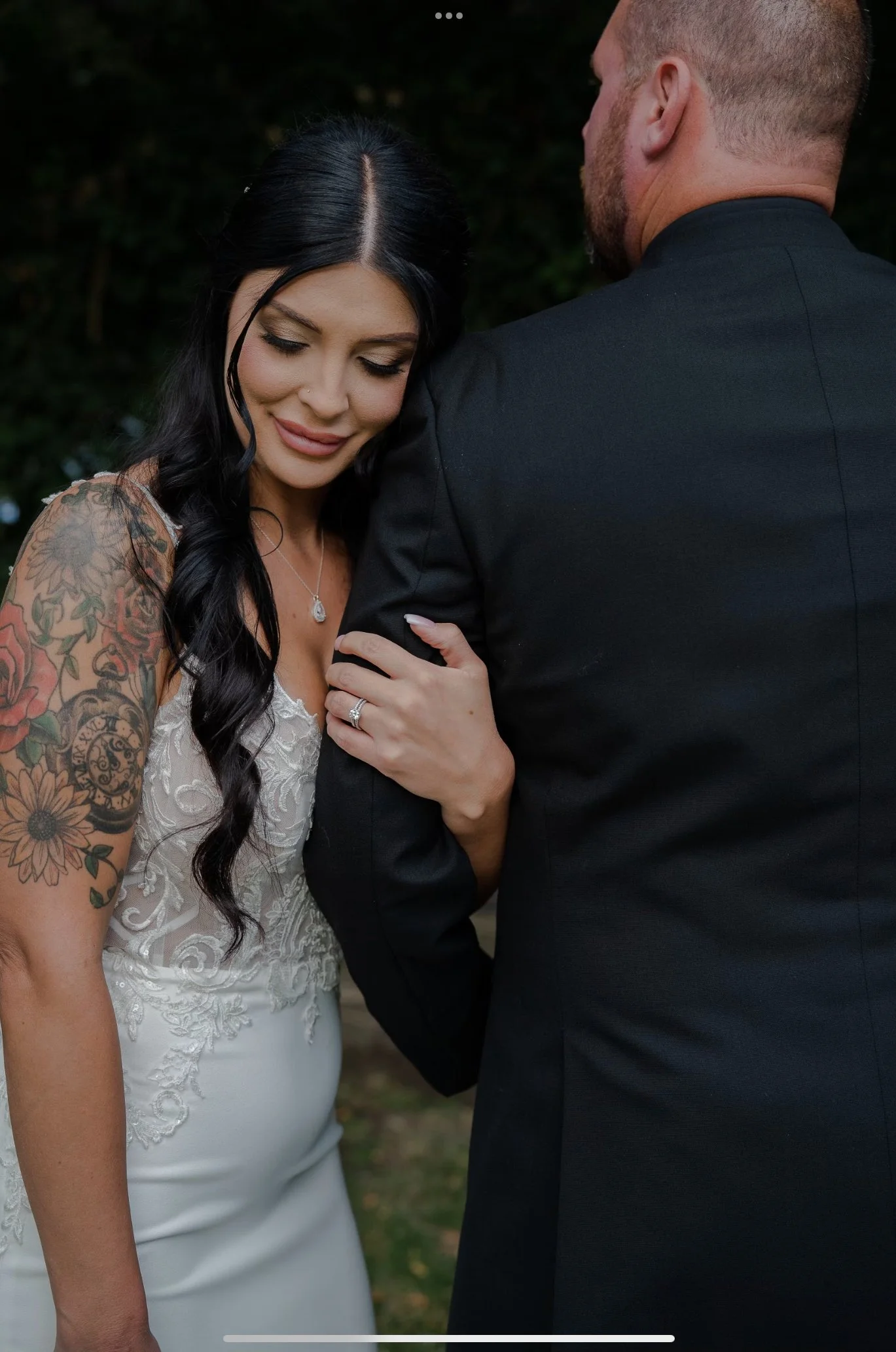 Close-up of a bride and groom on their wedding day, with the bride smiling and showing her wedding ring, while the groom is facing away from the camera. The bride has black hair, tattoos on her arm, and is wearing a lace wedding dress. The groom is d