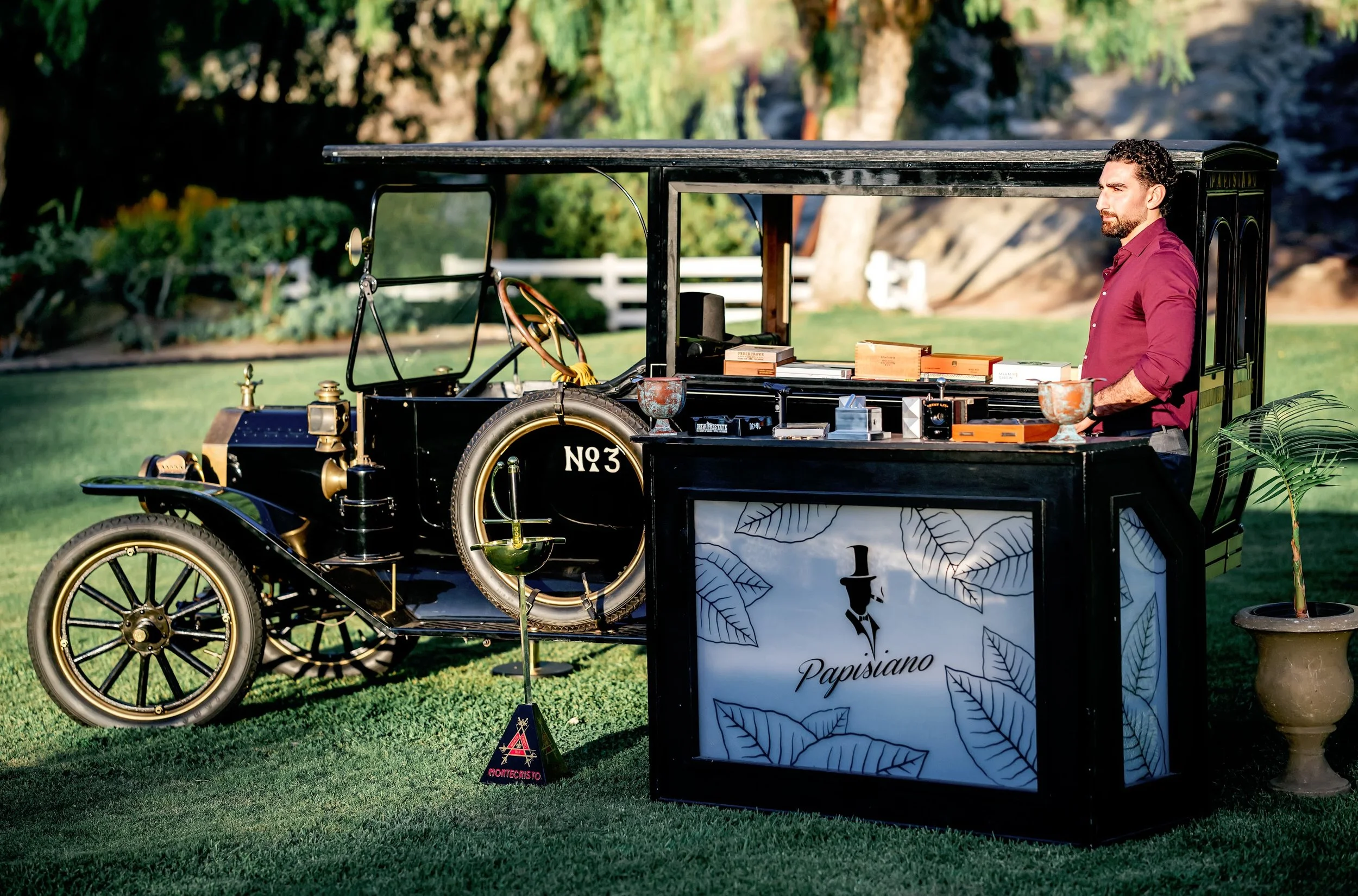A man in a maroon shirt stands next to a vintage black cart with decorative leaves and the name "Parsiano" printed on it. The cart displays books, small boxes, and decorative items, set outdoors on a grassy area with trees and a white fence in the ba