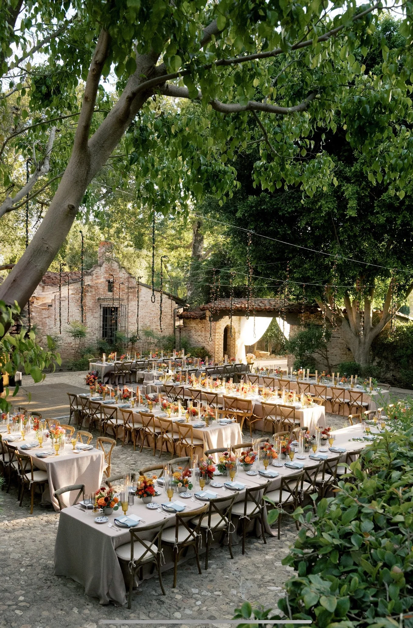 Outdoor wedding reception setup with long tables covered in beige tablecloths, adorned with floral centerpieces, set beneath large trees with hanging lights in a rustic courtyard.