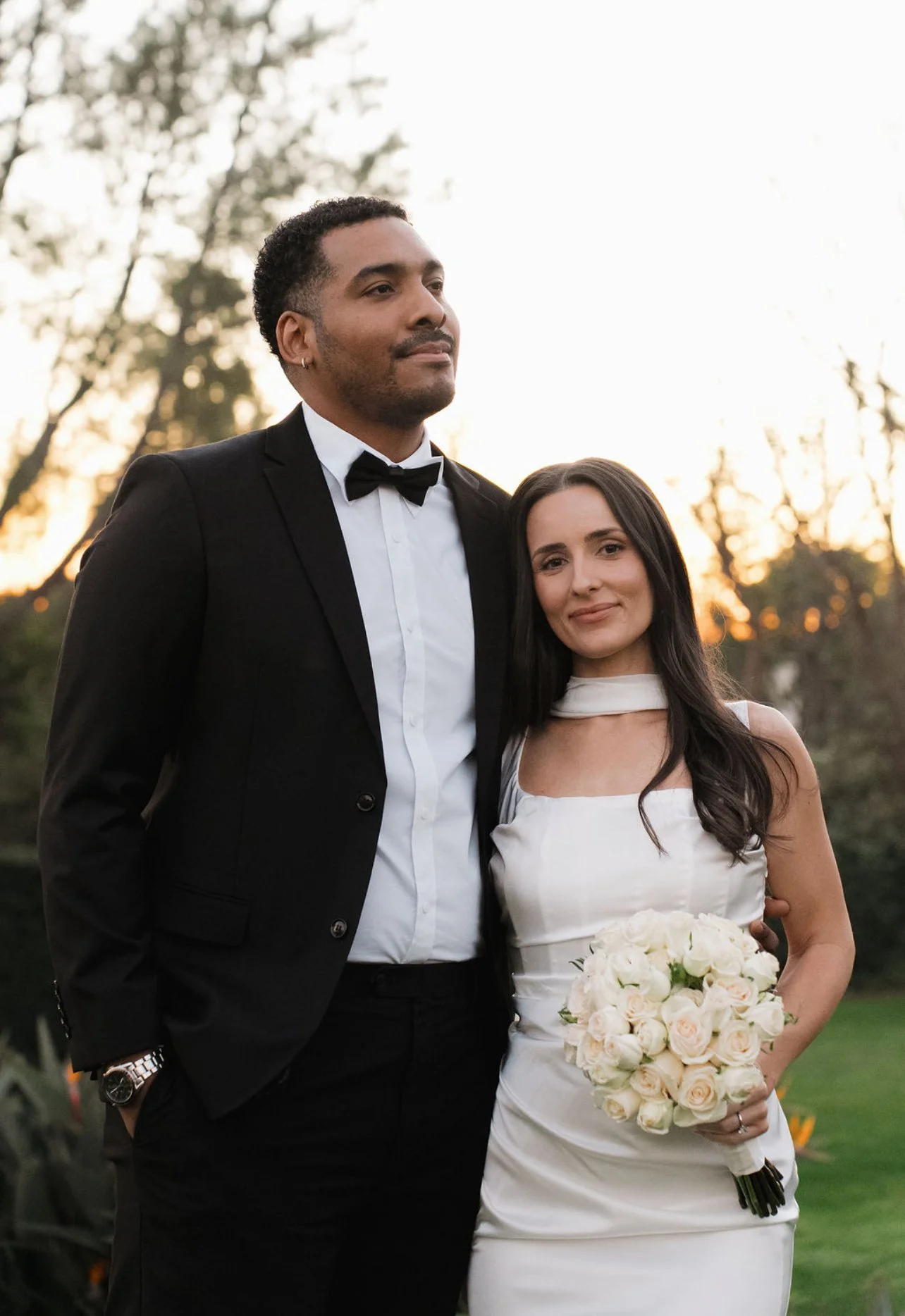 A newlywed couple outdoors at sunset, the groom in a black tuxedo and the bride in a white wedding dress holding a bouquet of white roses.