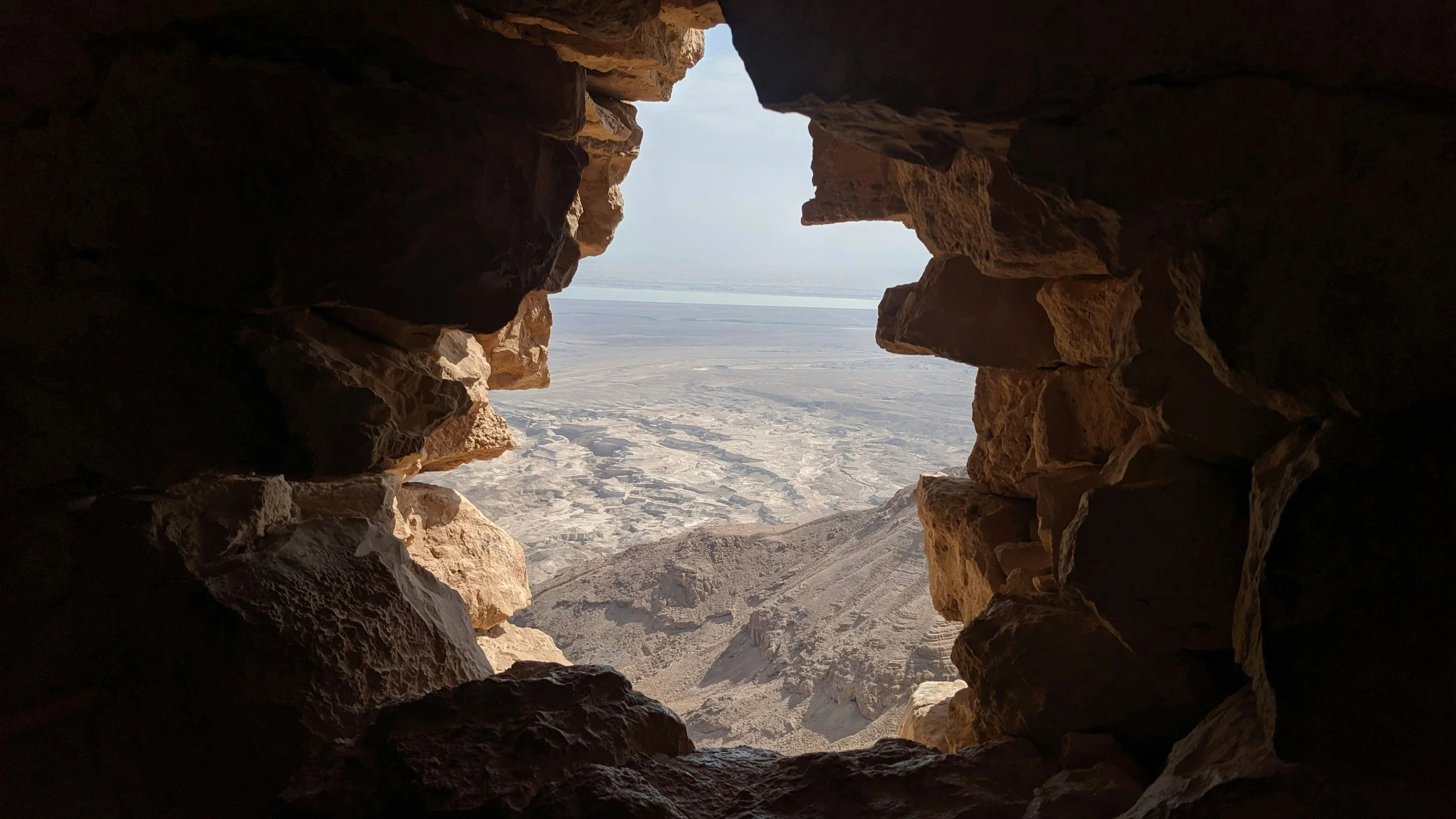 View through a small opening in a stone wall showing a desert landscape with hills and flat land in the distance.