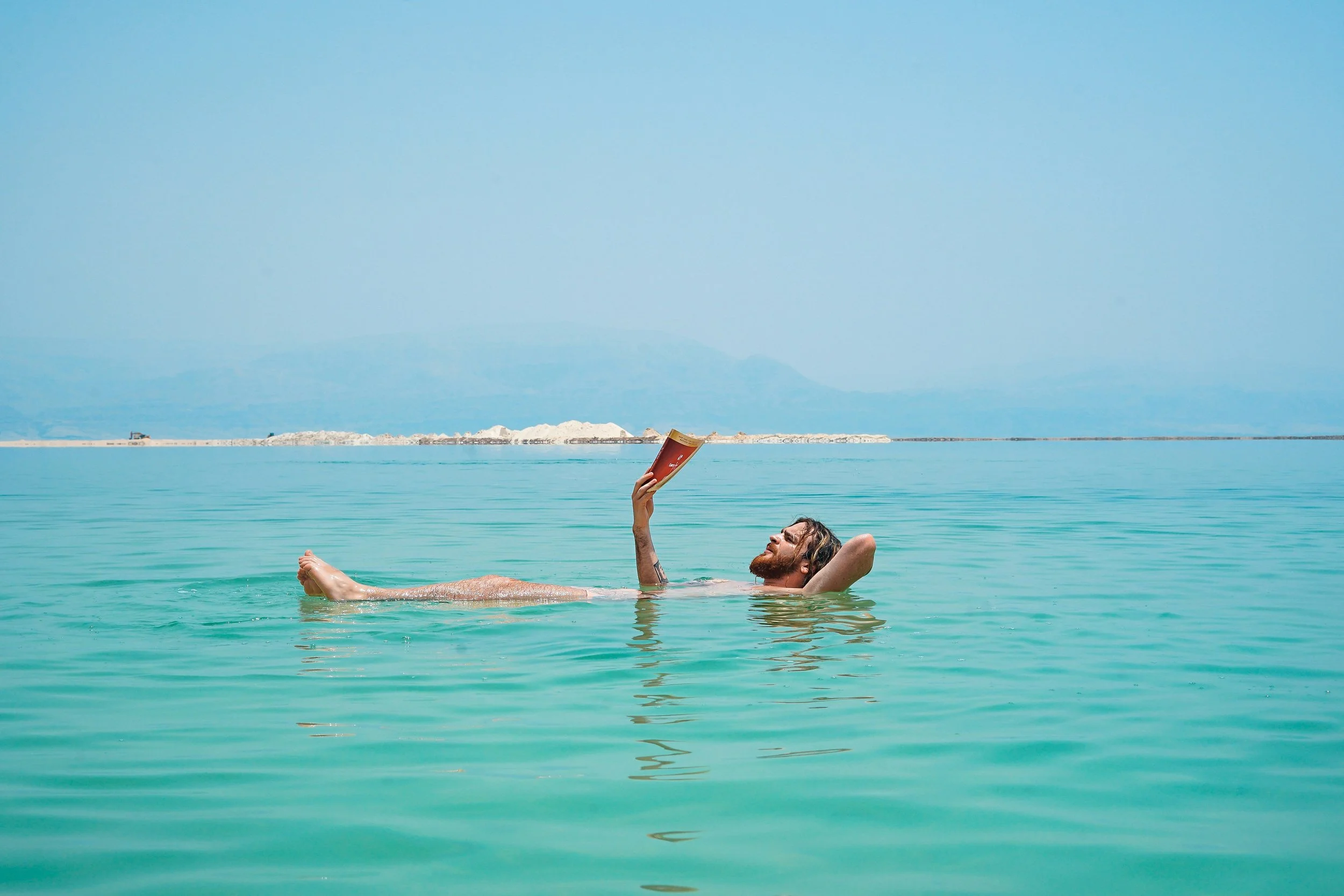 A man with a beard and long hair reading a book while floating on his back in calm turquoise water, with distant mountains and a clear blue sky in the background
