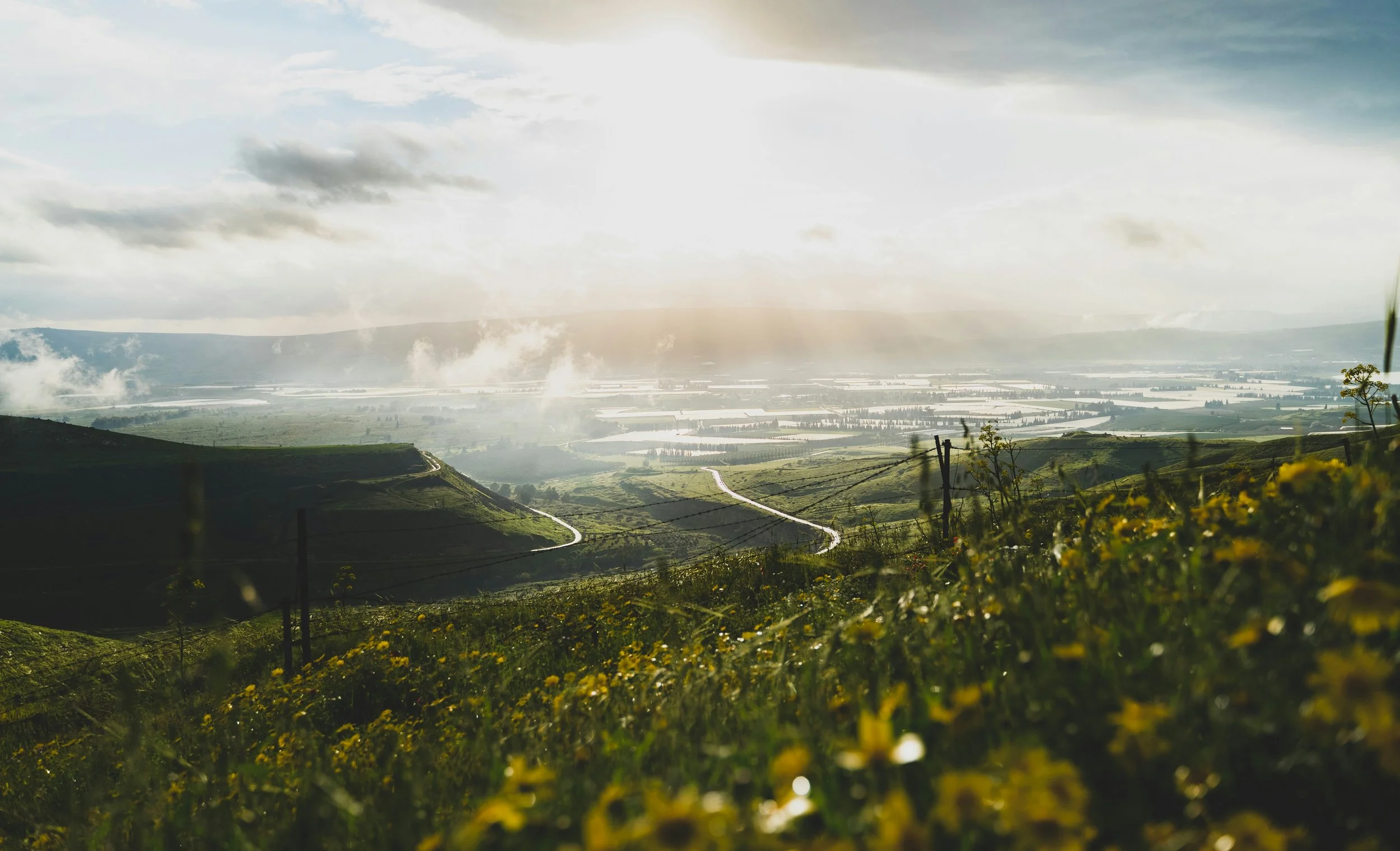 View of rolling green hills with a winding dirt road, power lines, and a vast valley with water bodies in the distance, under a partly cloudy sky with sunlight breaking through.