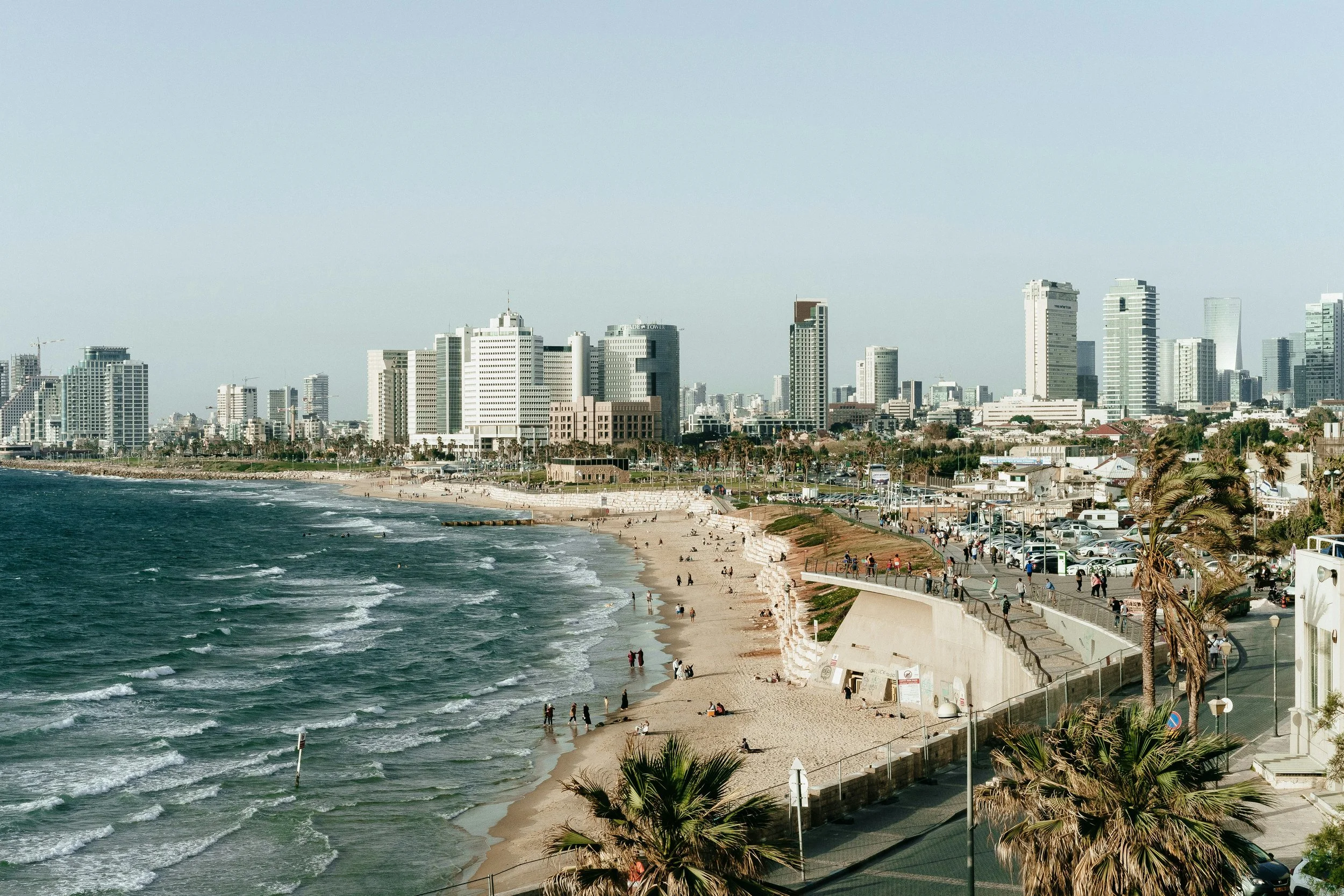 A city skyline with tall buildings along a coastline, a sandy beach with people walking and sitting, and palm trees lining a promenade.