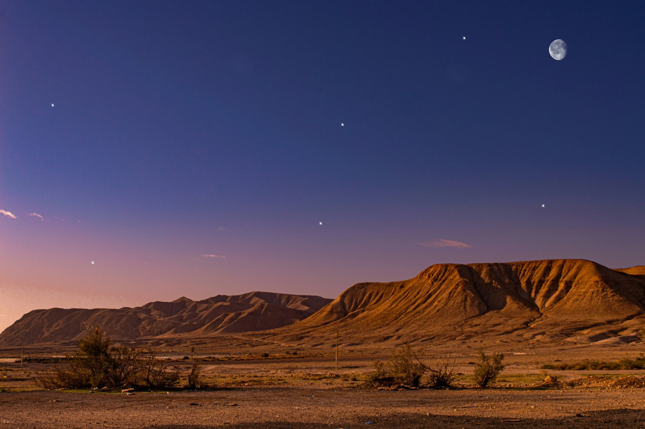 A nighttime desert landscape with mountain ranges under a dark sky, featuring visible stars, the moon, and a bright planet or star.