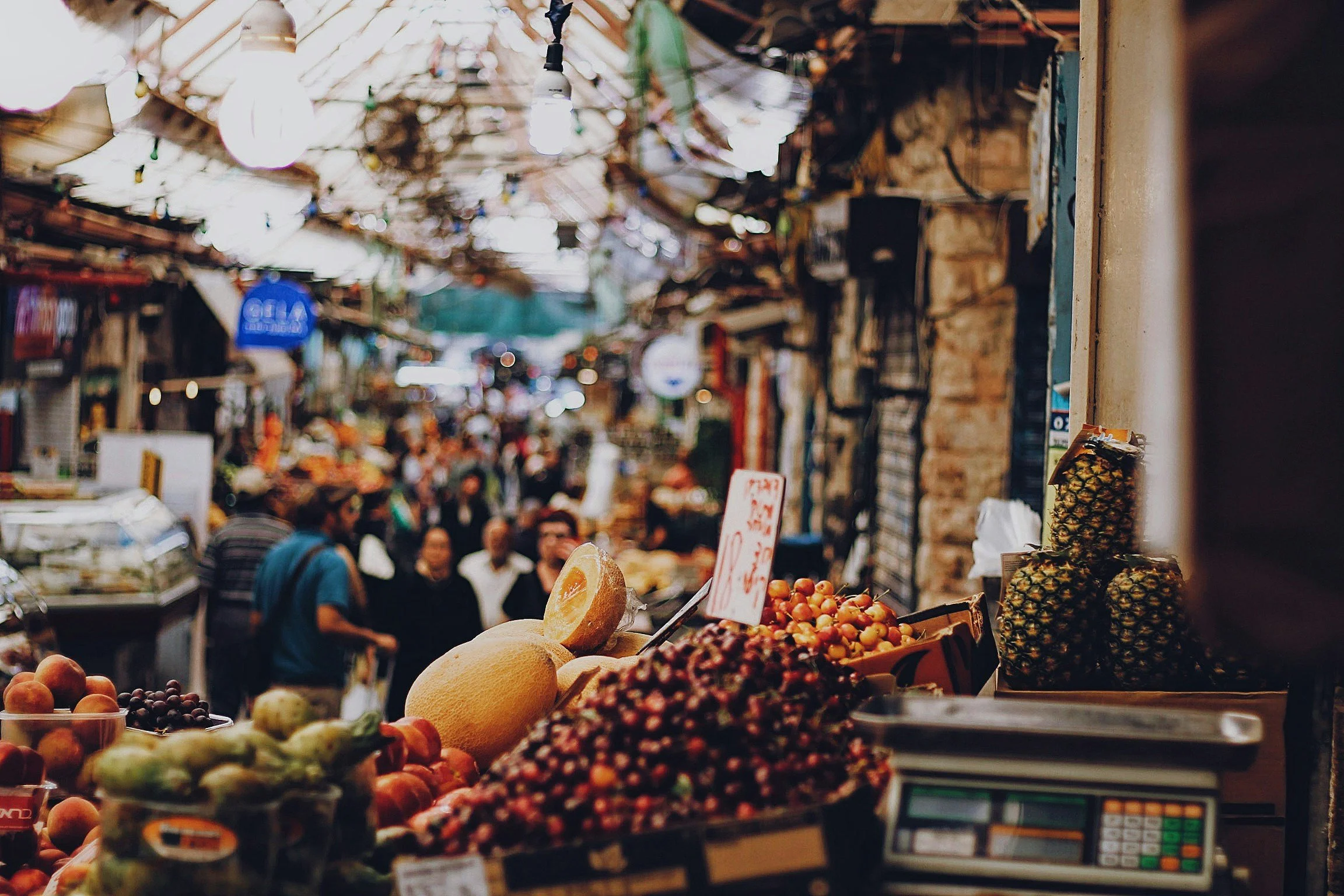 Fruits at a market stall, with melons, grapes, cherries, pineapple, and other produce, in a busy outdoor marketplace.