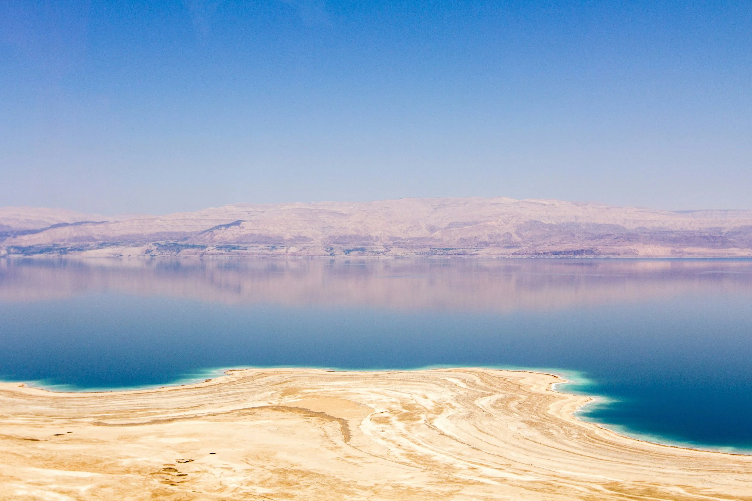 A large lake with clear, blue water reflecting the distant hills and sky, with a sandy shoreline in the foreground.