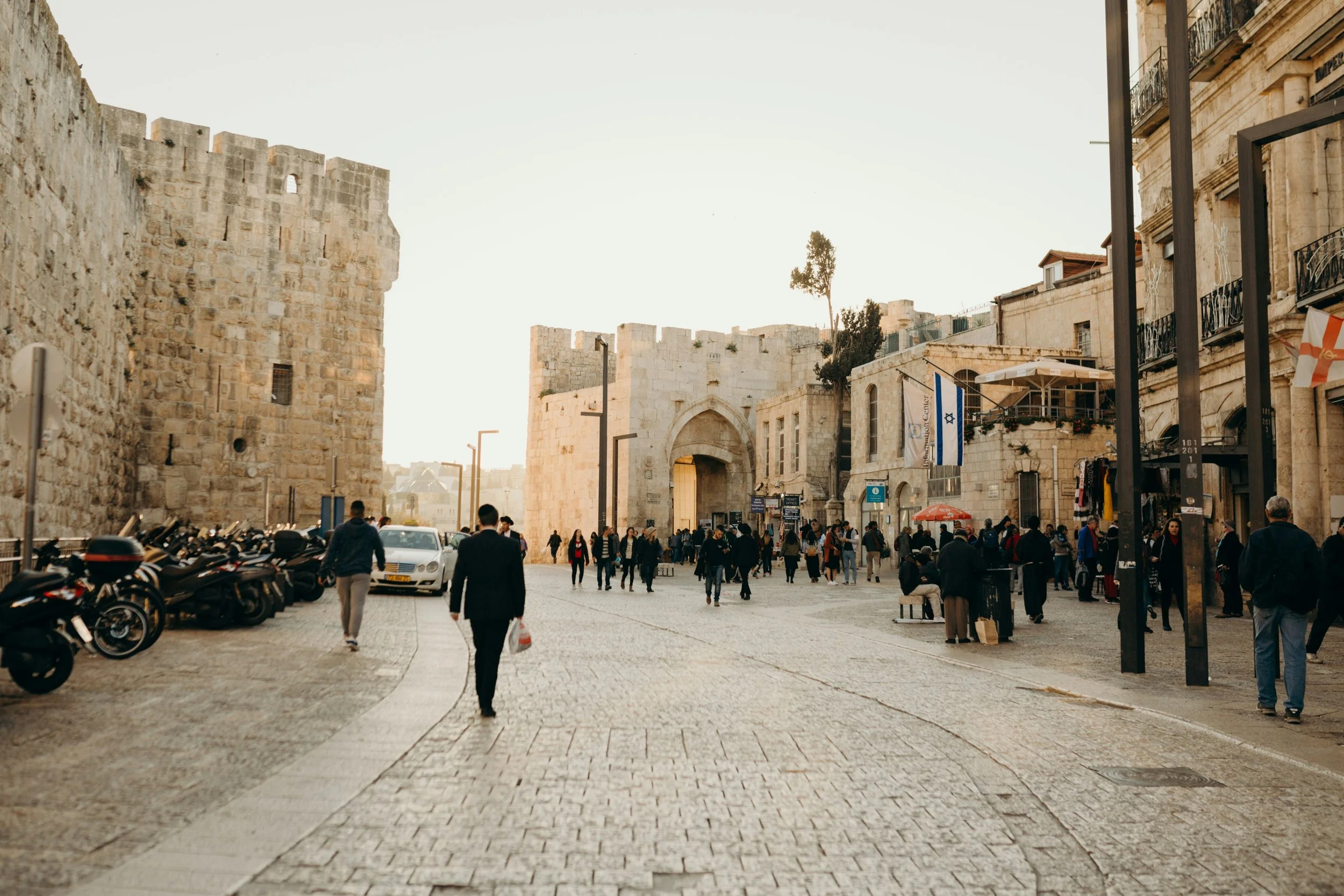 A busy city street with stone buildings and an arched gateway in the background. People are walking and shopping, with some parked motorcycles and a white car on the left. Flags are hanging from the buildings, and there are street lamps and benches.