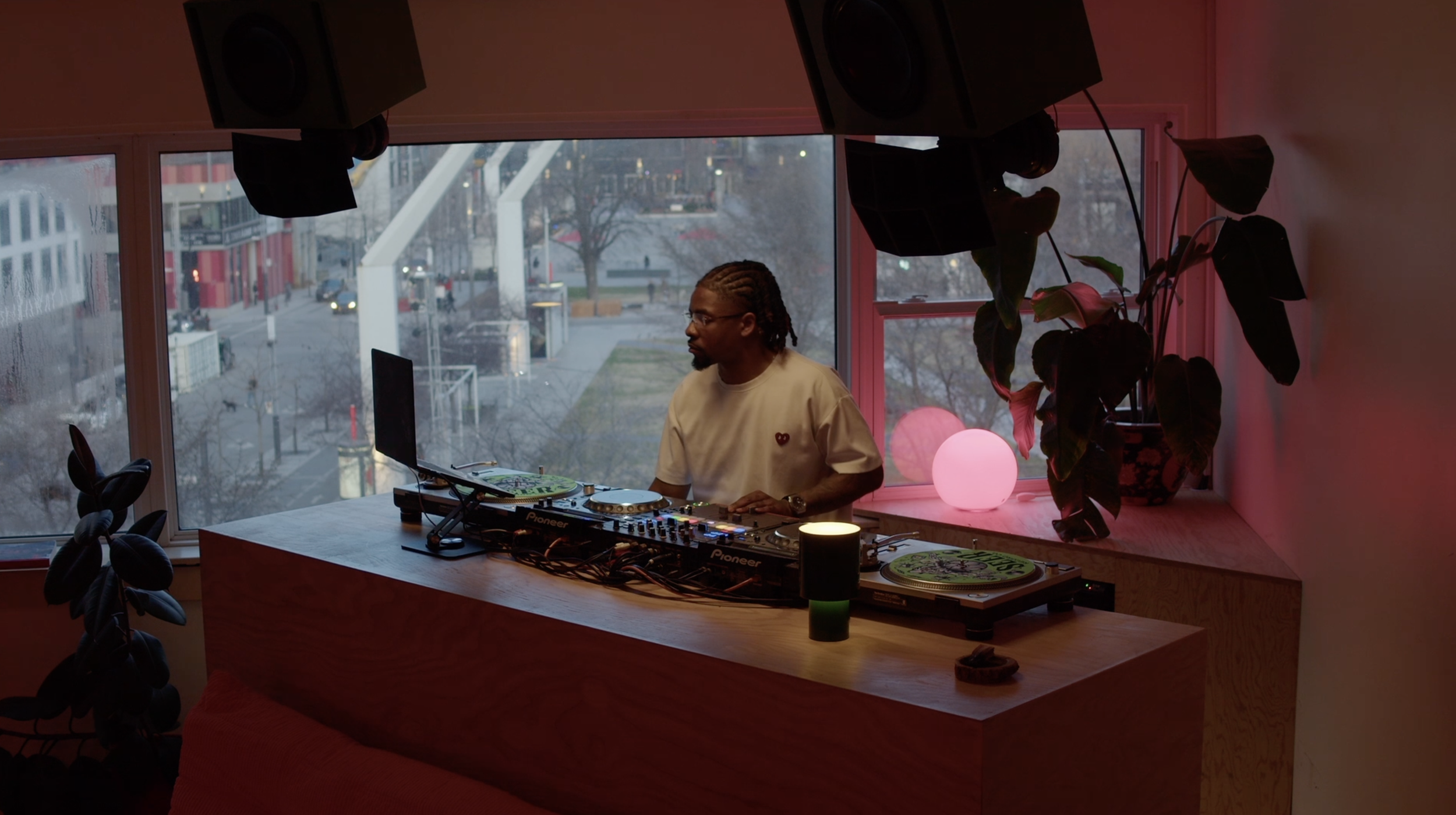 A man using DJ equipment stands in front of an apartment window overlooking Montreal