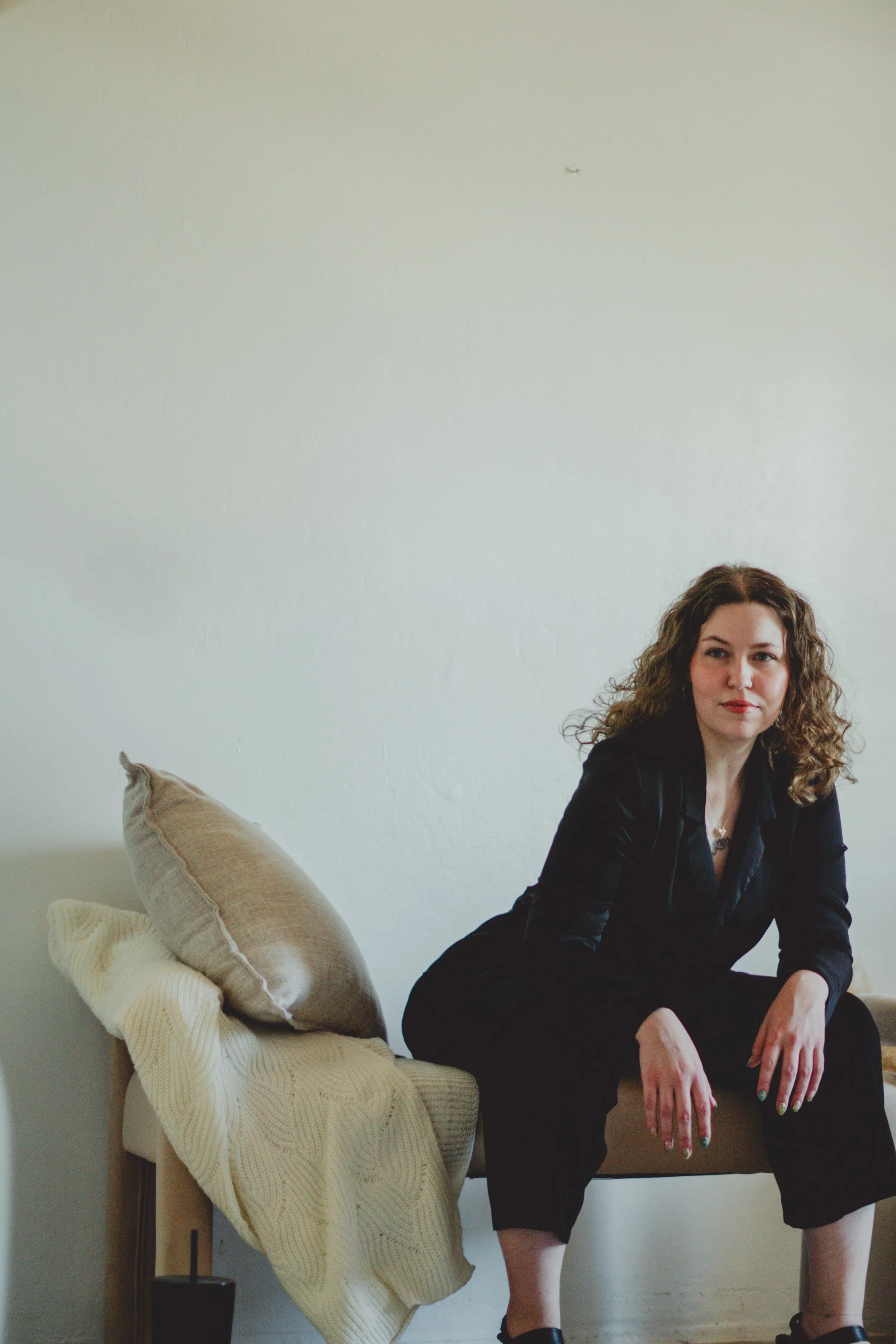 A woman with curly brown hair wearing a black outfit sitting on a beige bench with a pillow and a blanket in a room with a bare white wall.