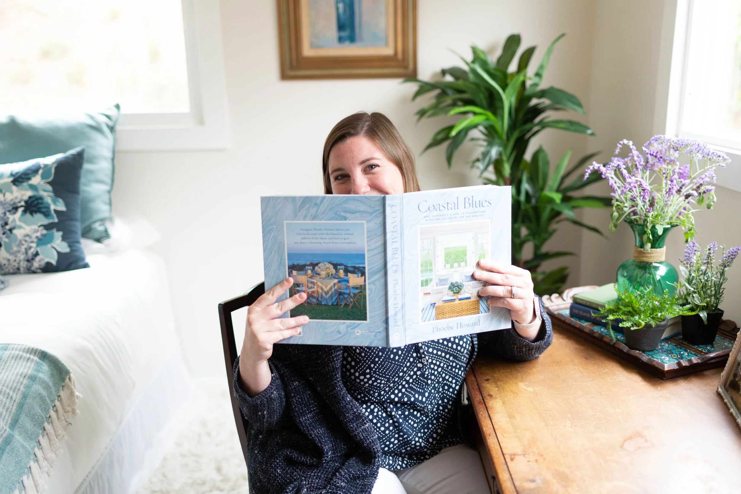 Christine Craig, owner of Designology Studio holding a book in a bedroom with natural light, indoor plants and wood furniture creating a relaxed biophilic design that feels calm and personal.