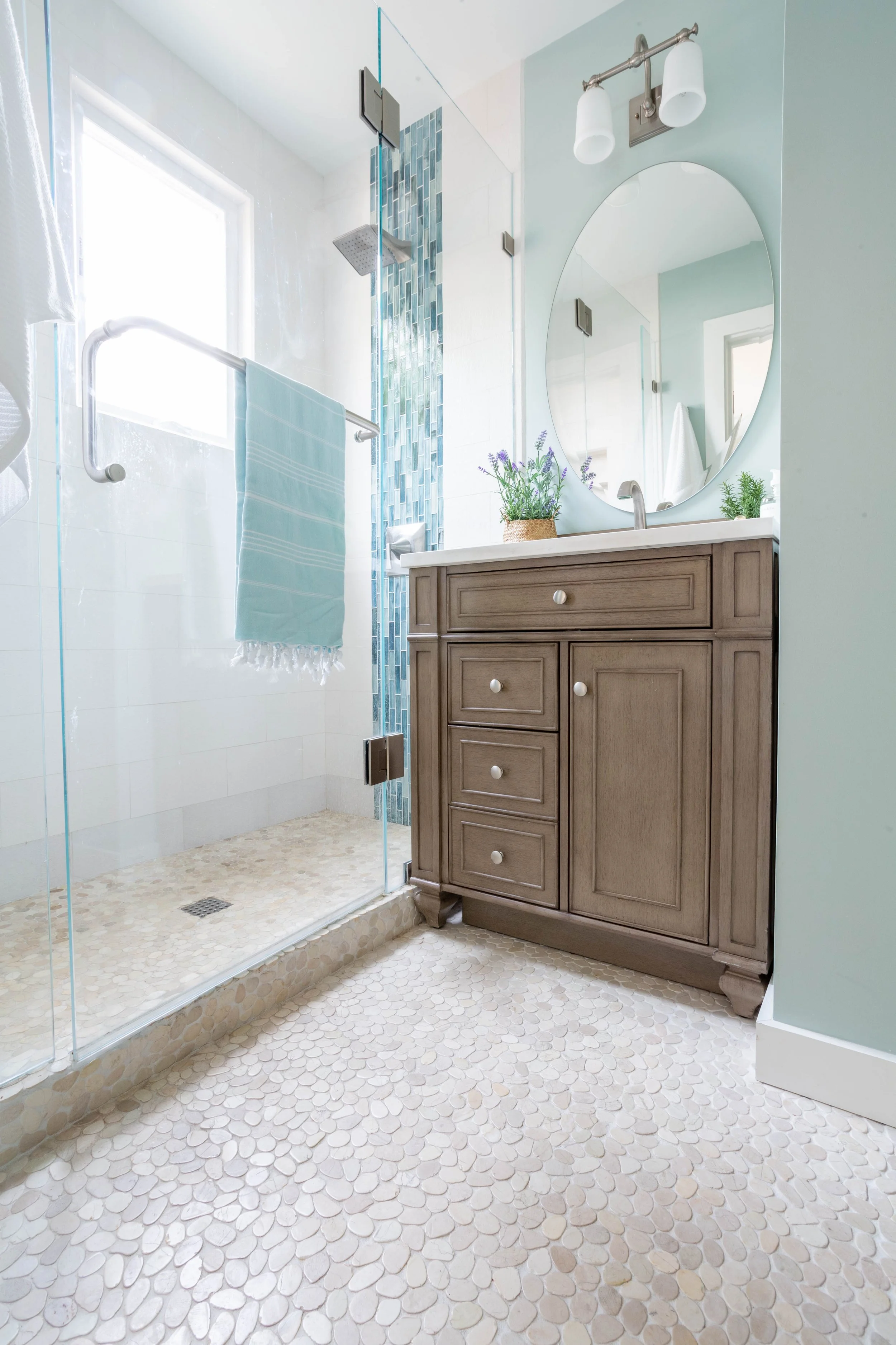 Bright primary bathroom with natural light, wood vanity, pebble tile floor, glass shower, and soft blue tones reflecting biophilic bathroom design focused on calm and wellness.