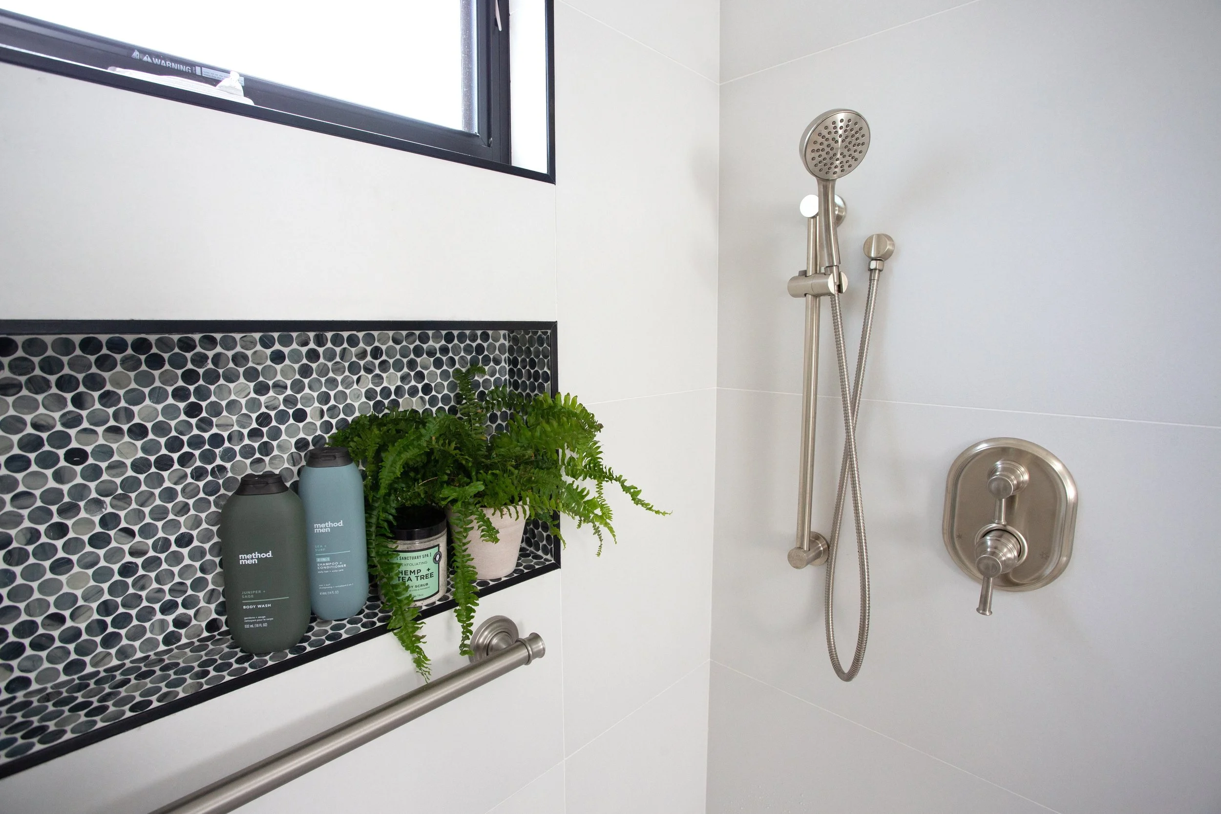 Curbless shower with built-in niche, mosaic tile accent, handheld shower head, and integrated grab bar for safe, stylish aging-in-place bathroom design.