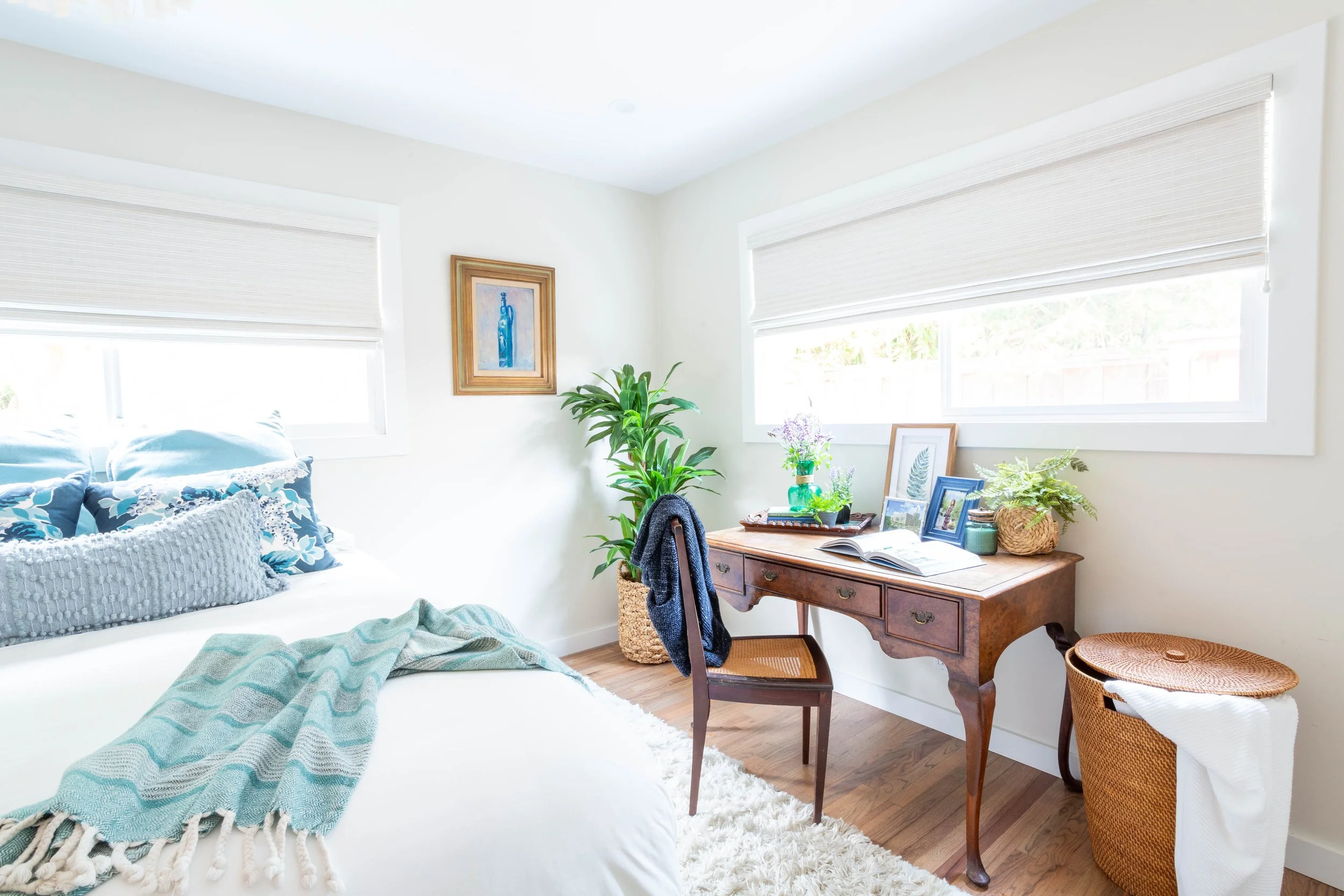 Bedroom with vintage writing desk, woven chair, plants, and layered bedding showing how heirloom furniture can elevate a space.
