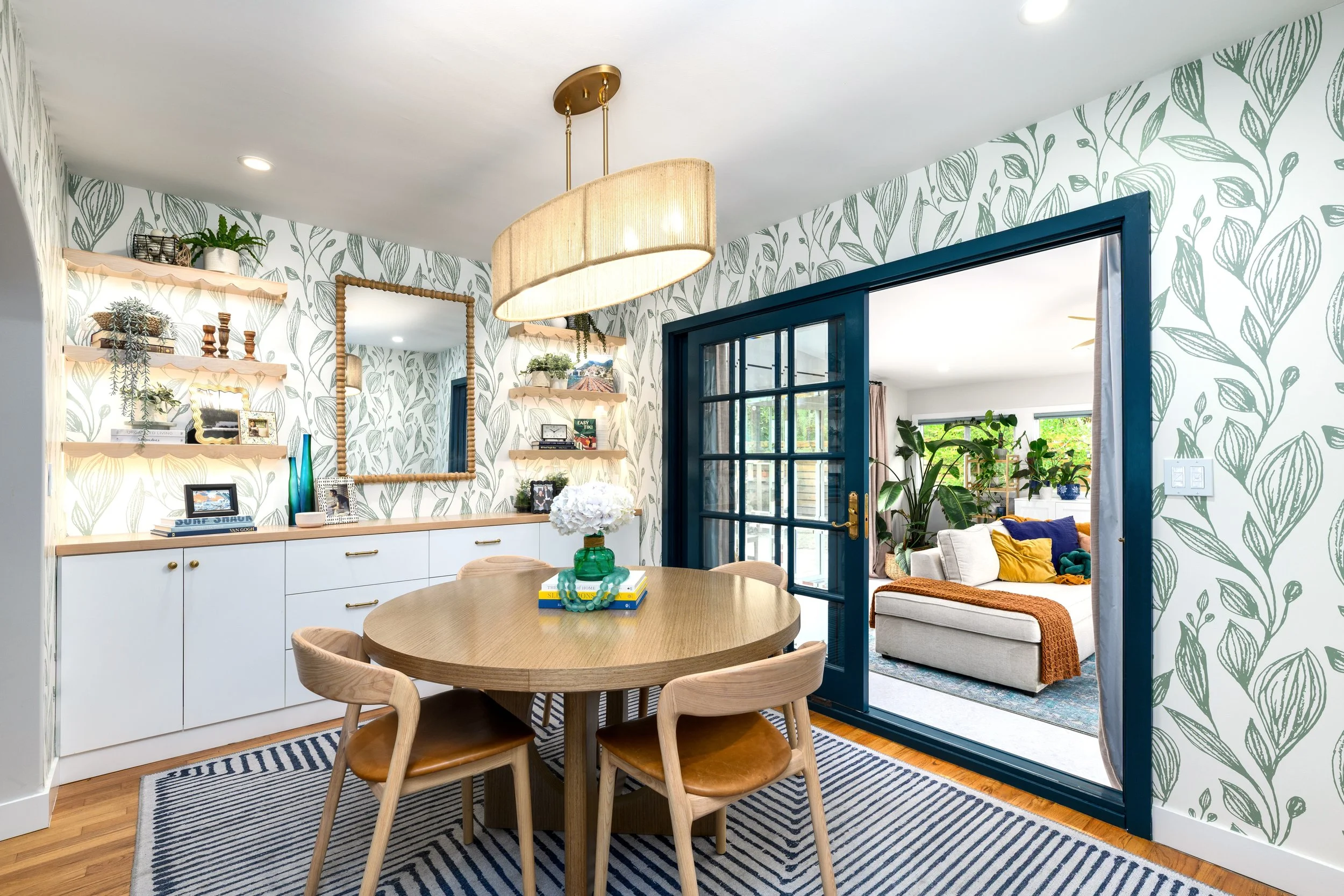 Dining room with botanical wallpaper, wood dining table, layered lighting, and view into a plant filled living room in a Ventura home.
