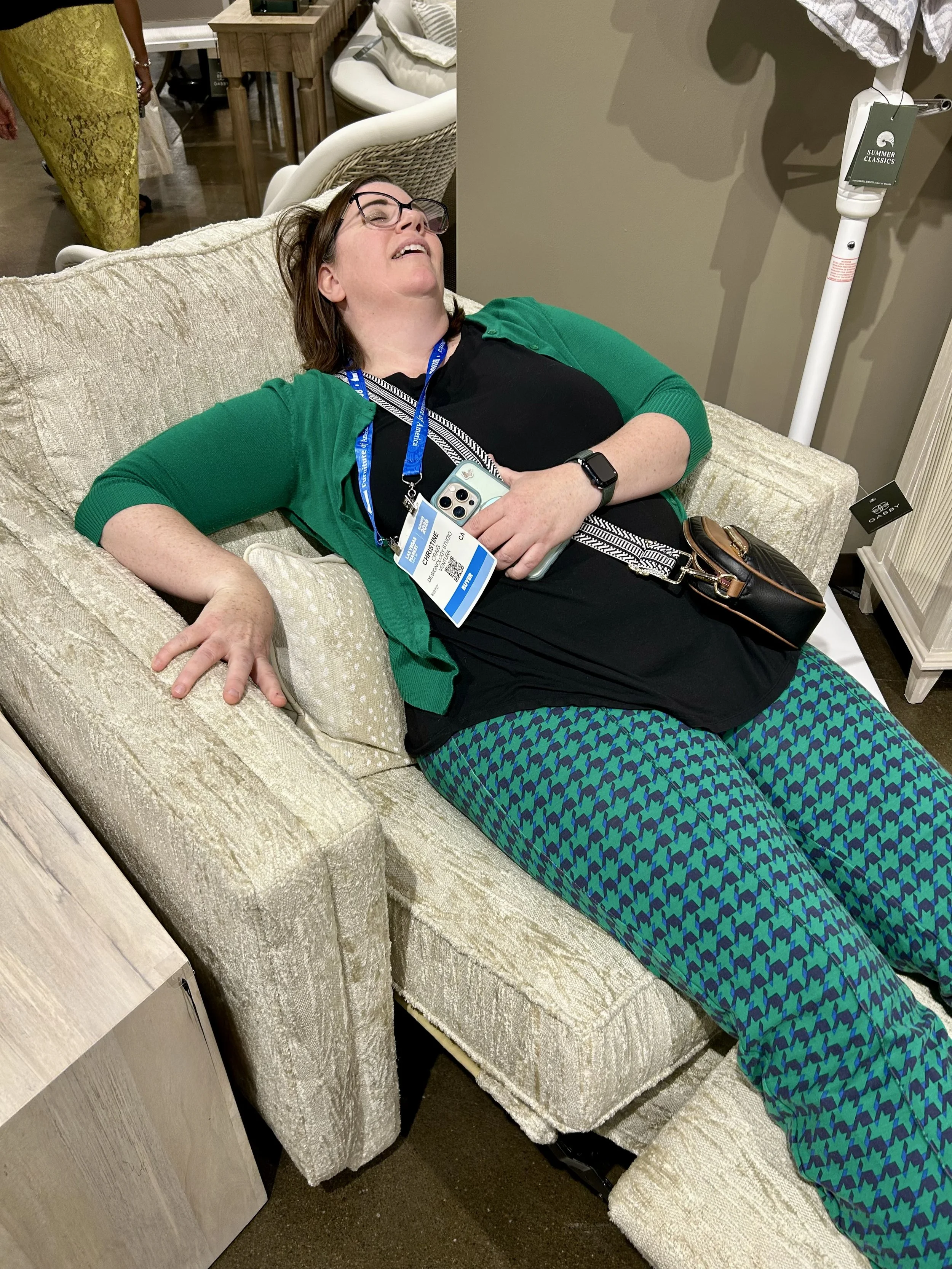 Designology Studio designer testing the comfort of an upholstered chair while sourcing furniture at Las Vegas Market.