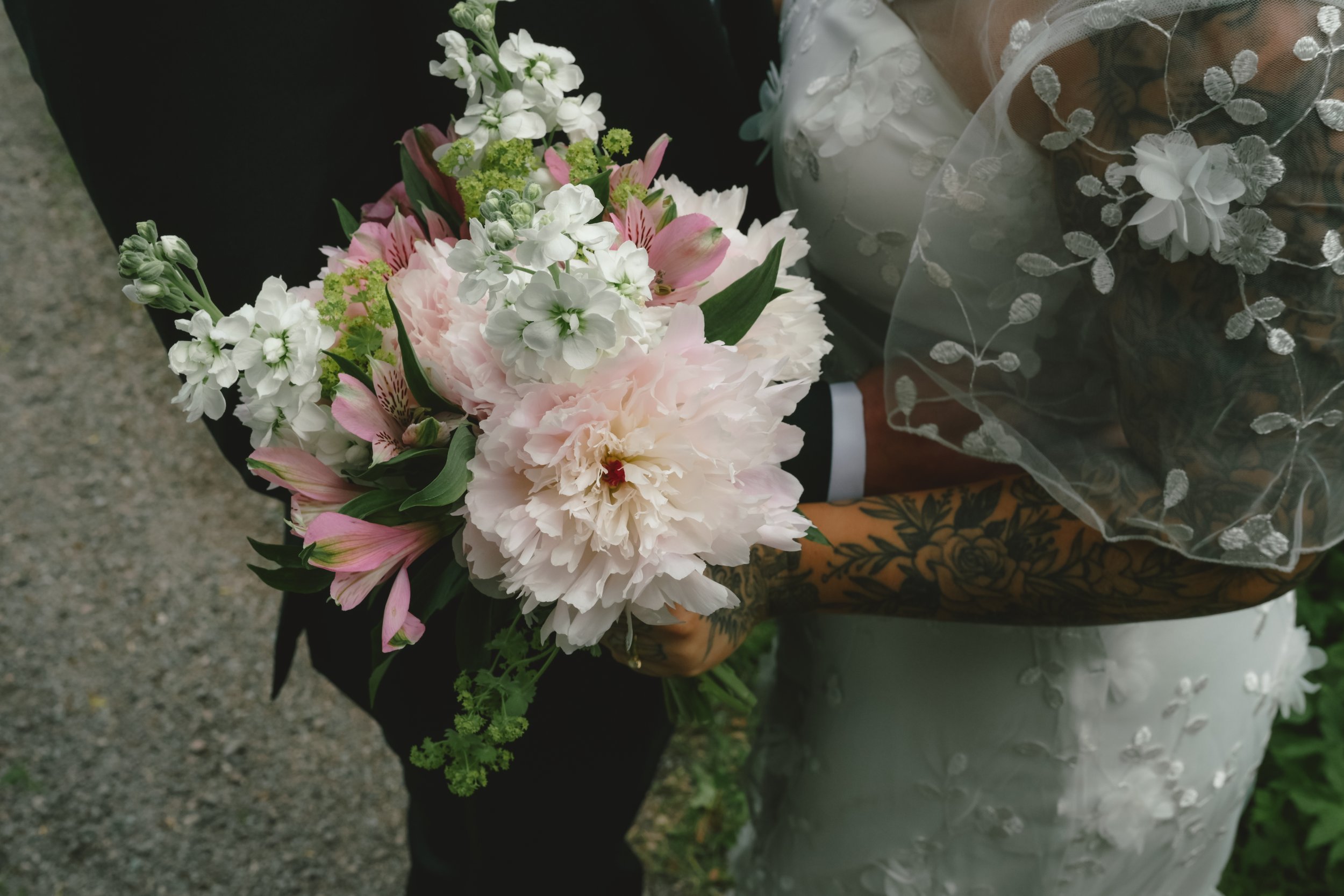 A bride holding a bouquet of pink and white flowers, including peonies, lilies, and small white blossoms, with the bride wearing a wedding dress with embroidered floral details and a sheer embroidered veil.