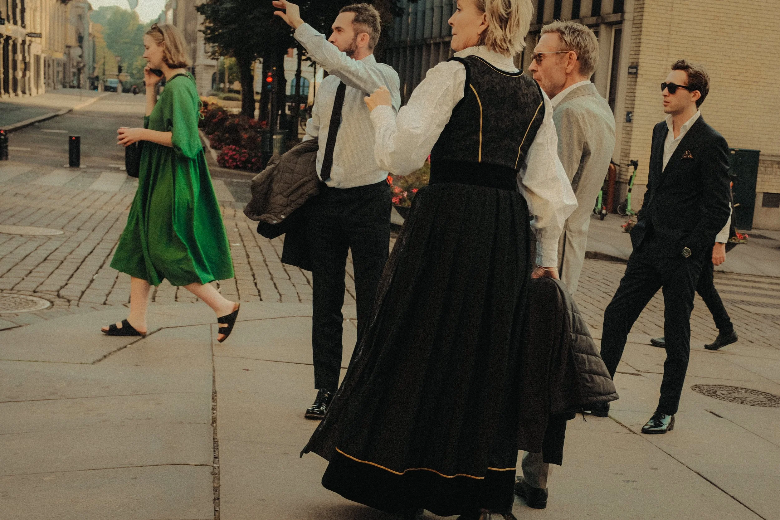 A group of people walking across a city street, with some engaged in conversations and one woman in a green dress talking on her phone.
