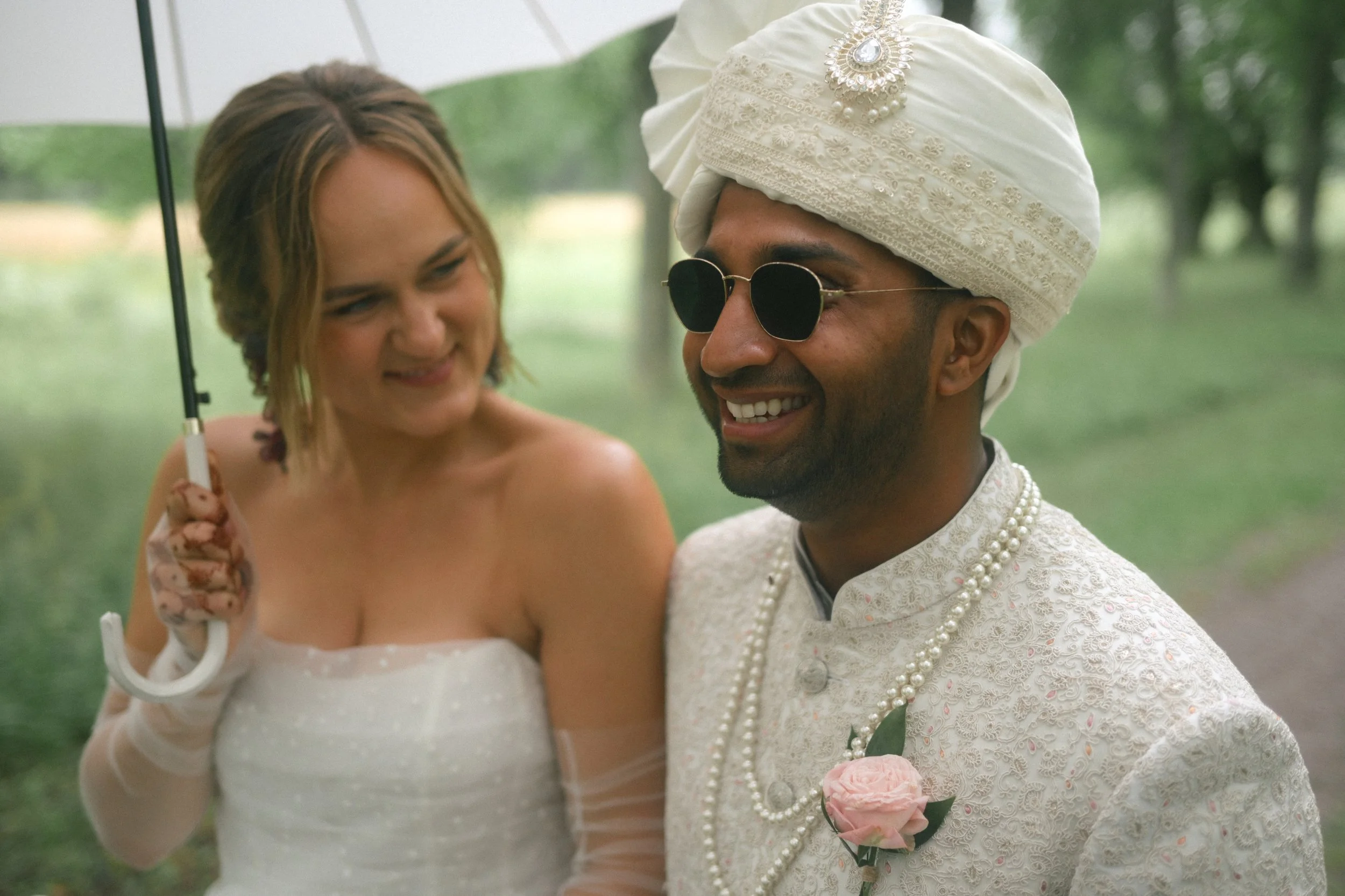 A bride and groom dressed in traditional Indian attire outdoors, with the bride holding an umbrella and the groom wearing sunglasses and a turban, smiling.