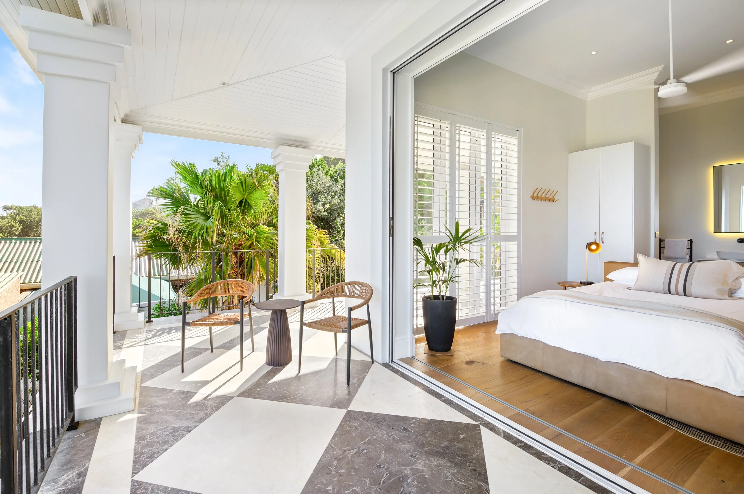 A balcony connected to a bedroom with white walls and large windows with shutters, adorned with a potted plant, and outdoor furniture including two chairs and a small table, overlooking trees.