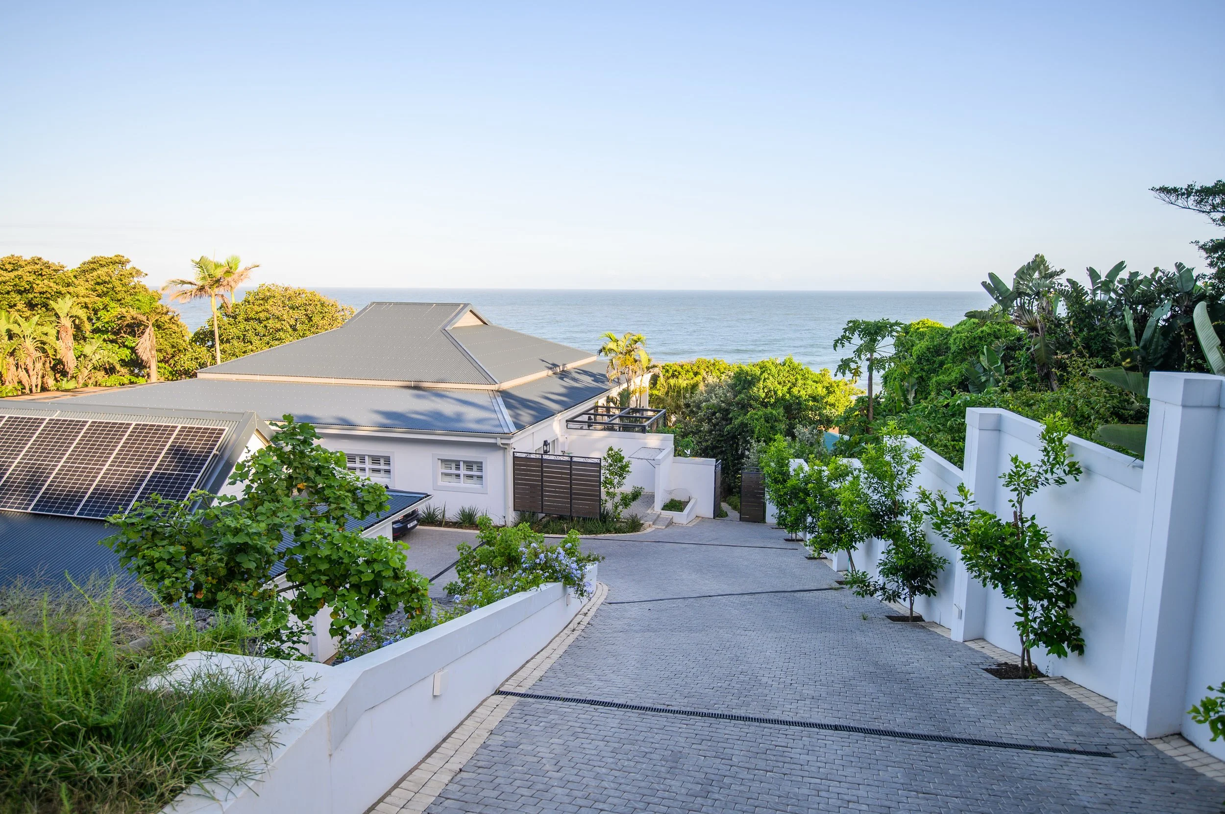 View of a residential property with a paved driveway, white walls, green trees, and a house with a gray roof, overlooking the ocean in the background.