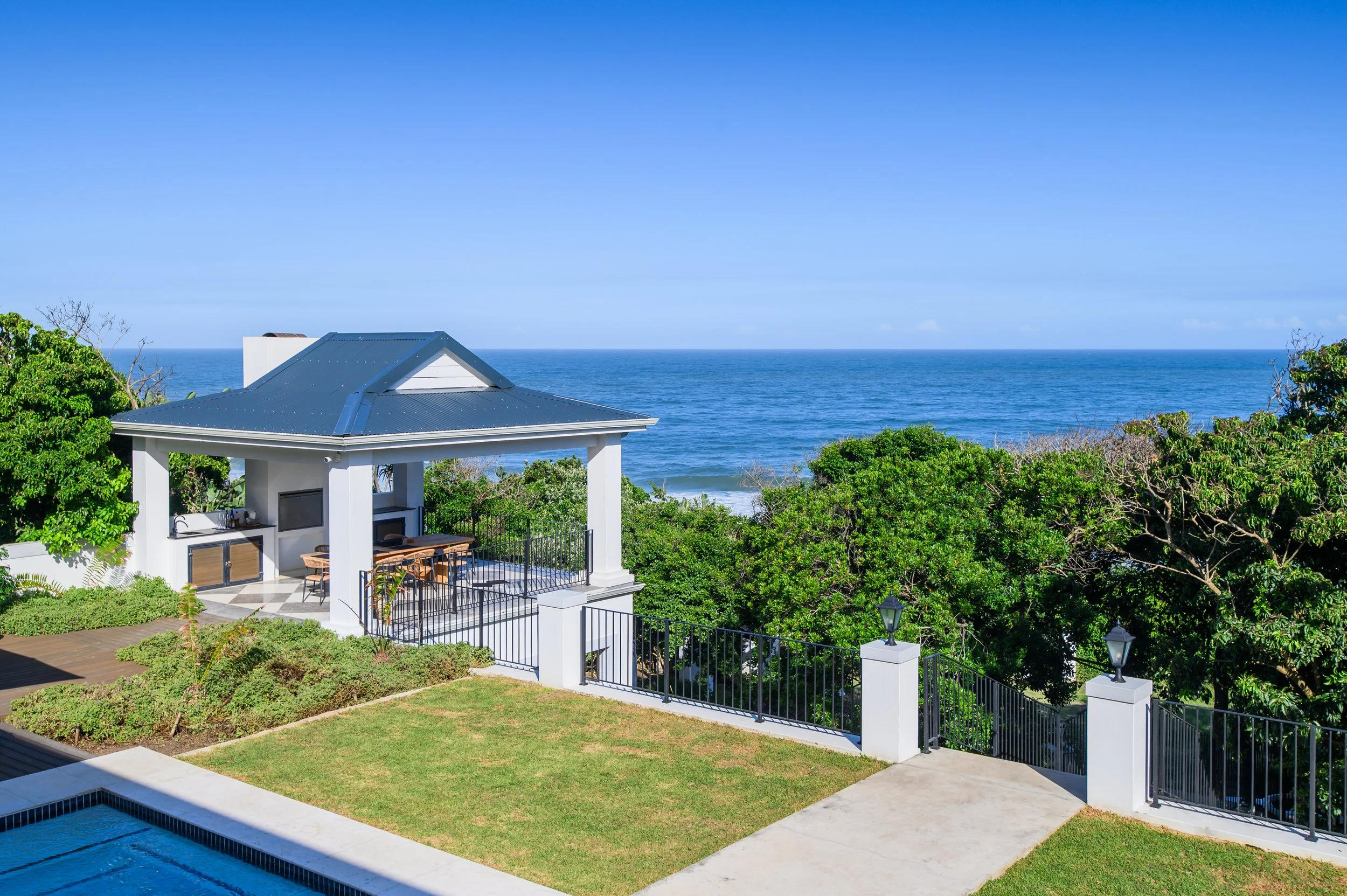 A house with a blue metal roof, an outdoor kitchen area, and a patio overlooking the ocean, surrounded by green trees and a fence.