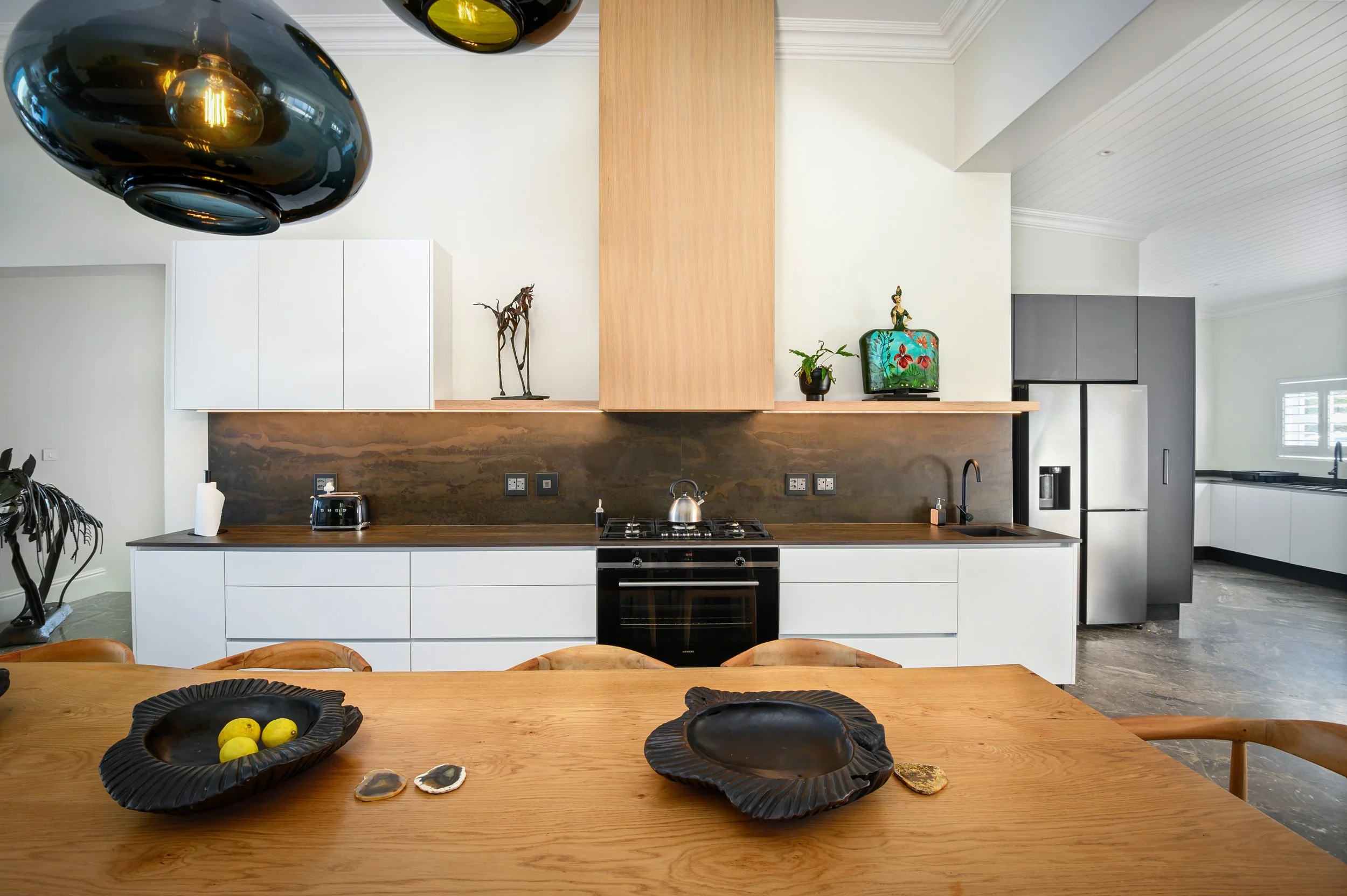Modern kitchen with white cabinets, a black stove, and a brown backsplash. Wooden dining table with black decorative bowls and lemons, and hanging black pendant lights.
