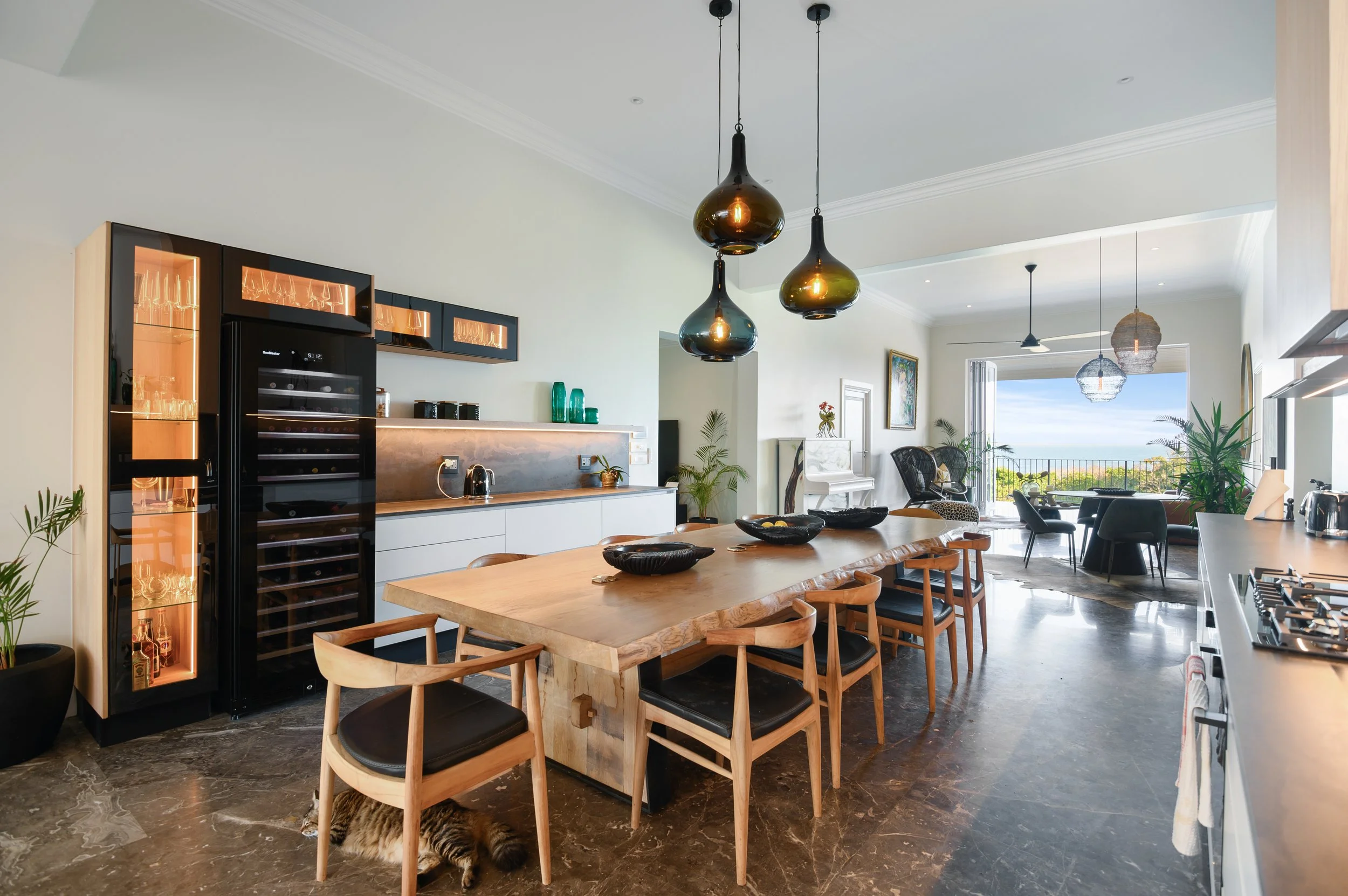 Open-concept kitchen and living room with a large wooden dining table, black pendant lights, a wine fridge, and a view of the balcony and ocean beyond.