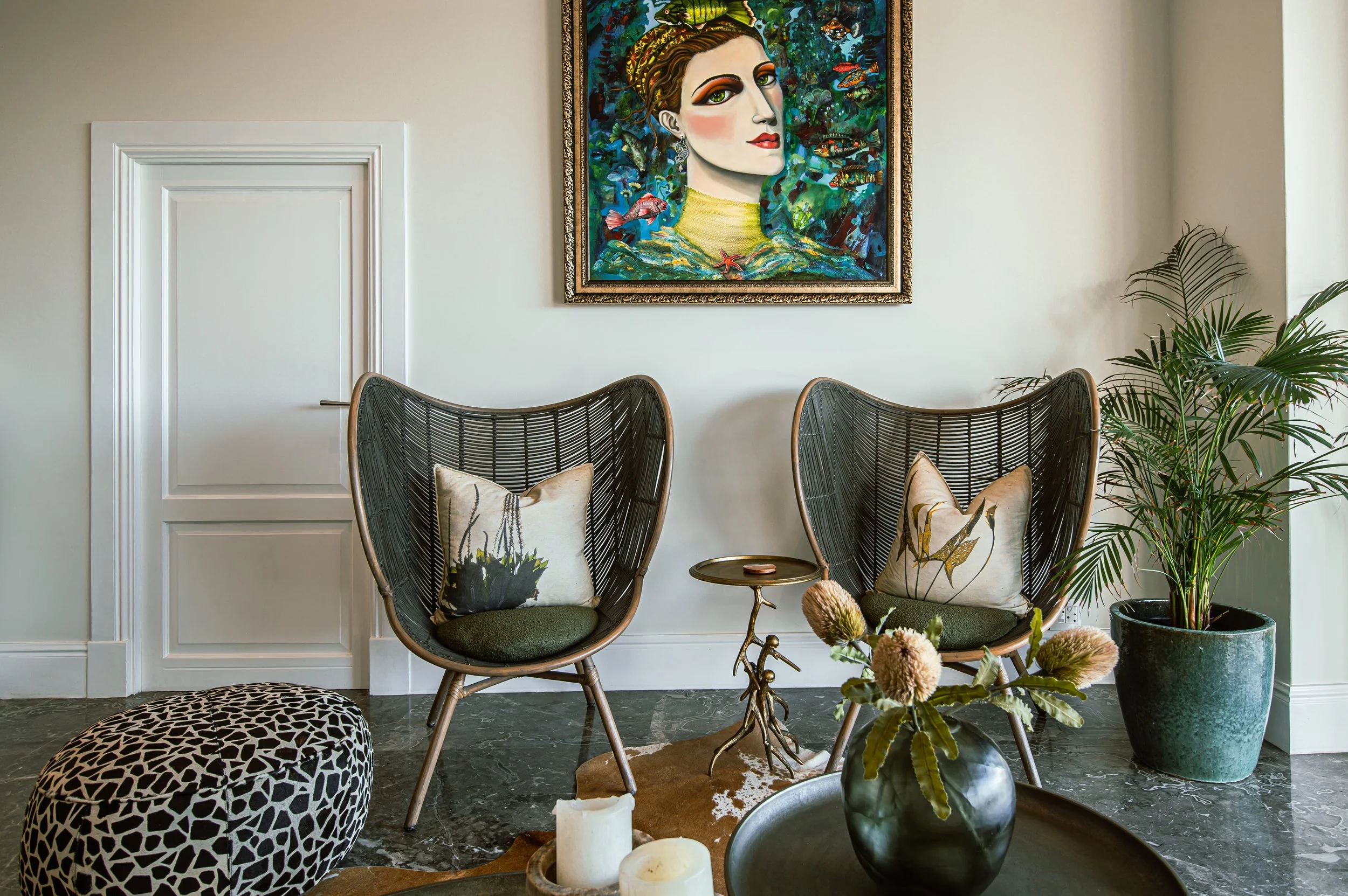 A cozy sitting area with two high-backed rattan chairs, each with a decorative pillow. Between them is a small side table with a branch-like design. To the right is a large potted palm plant. In front is a dark round table with a black vase holding f