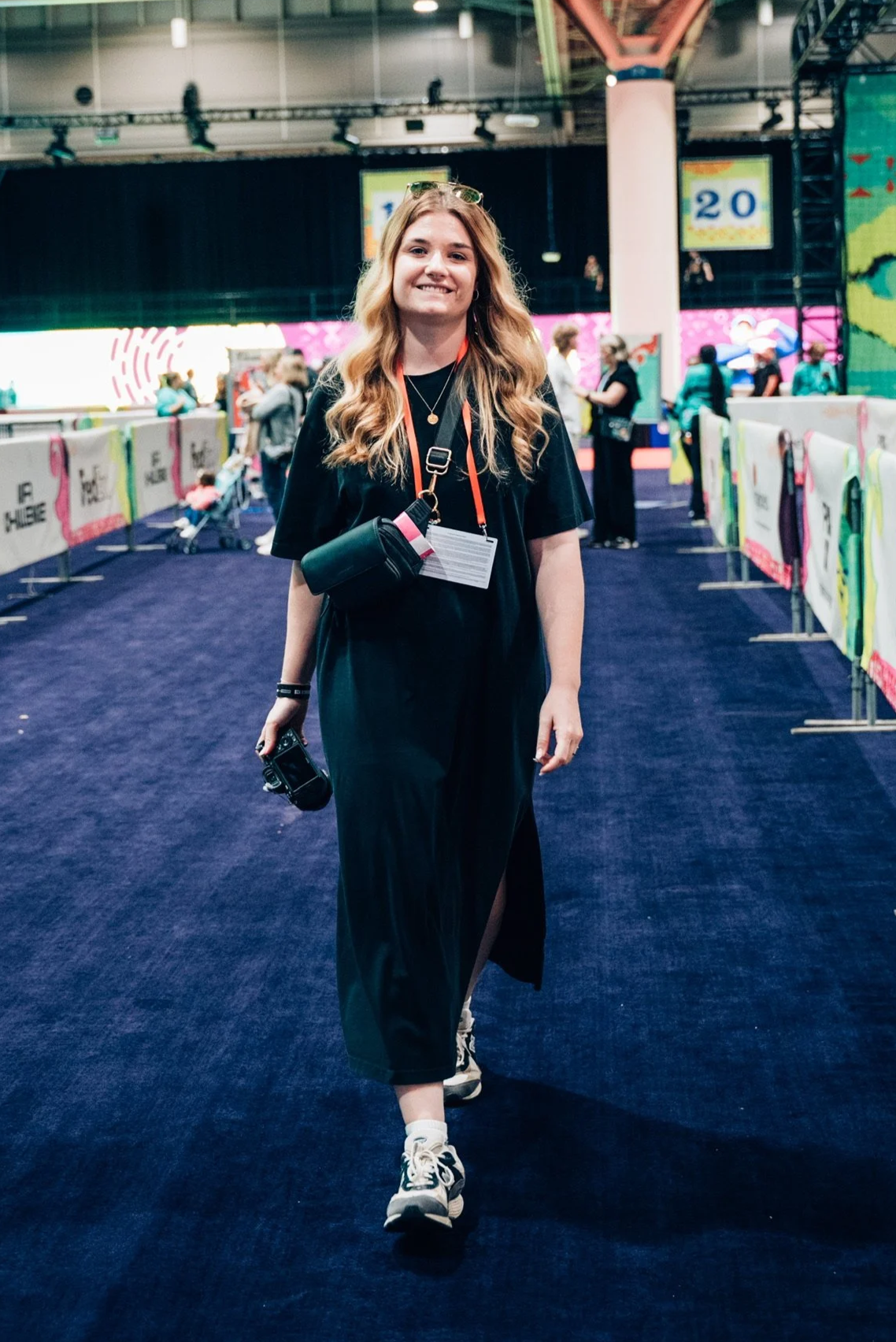 Eine junge Frau in einem schwarzen Kleid und Turnschuhen läuft in der NFL Experience in Las Vegas. Sie trägt Sonnenbrillen auf dem Kopf und hält eine Kamera in der Hand. Im Hintergrund sind mehrere Menschen und bunte Absperrgitter.
